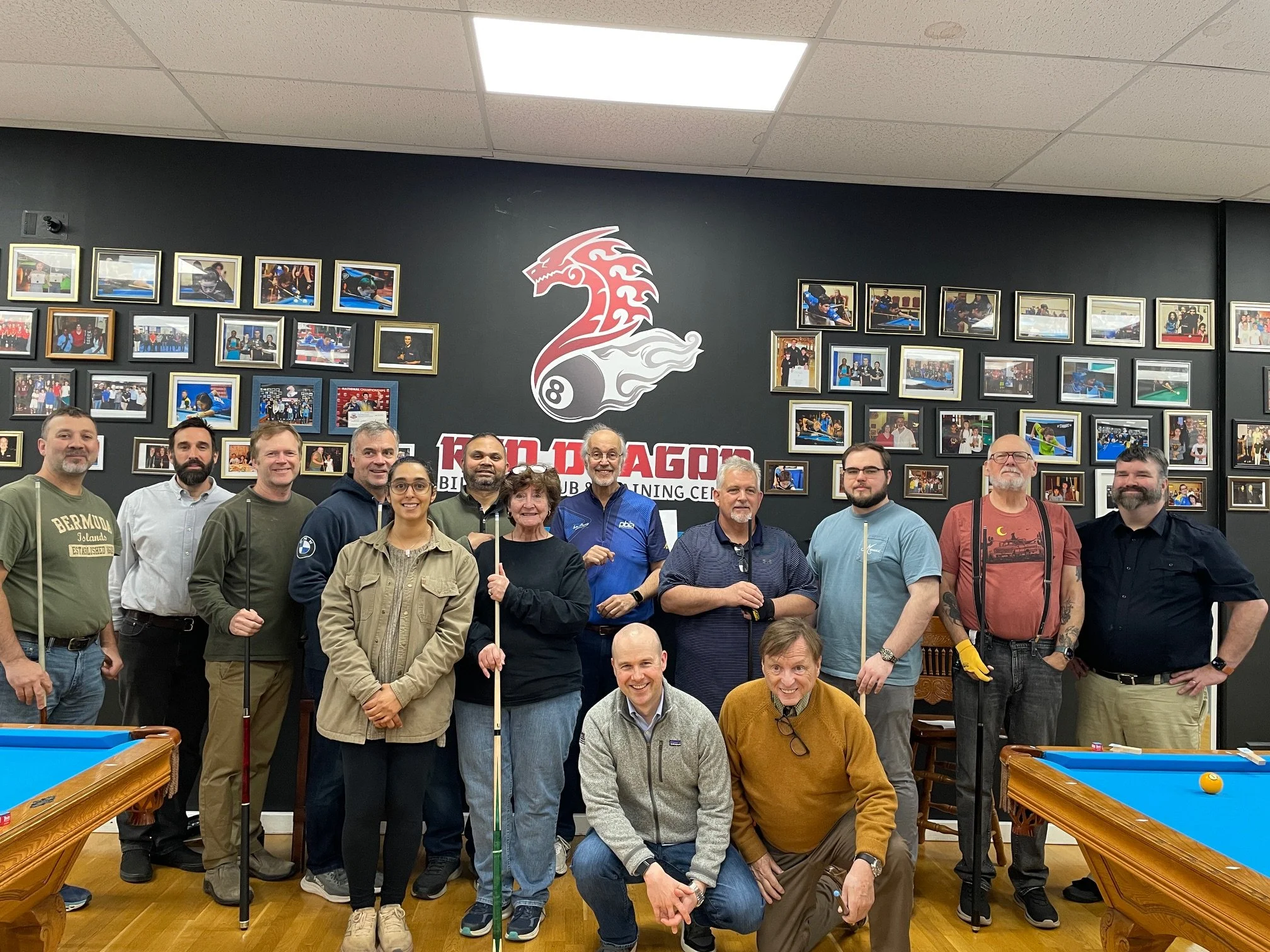 A group of people standing together in a billiards room, with a wall decorated with framed photos and a large logo of a dragon and an eight ball in the background.