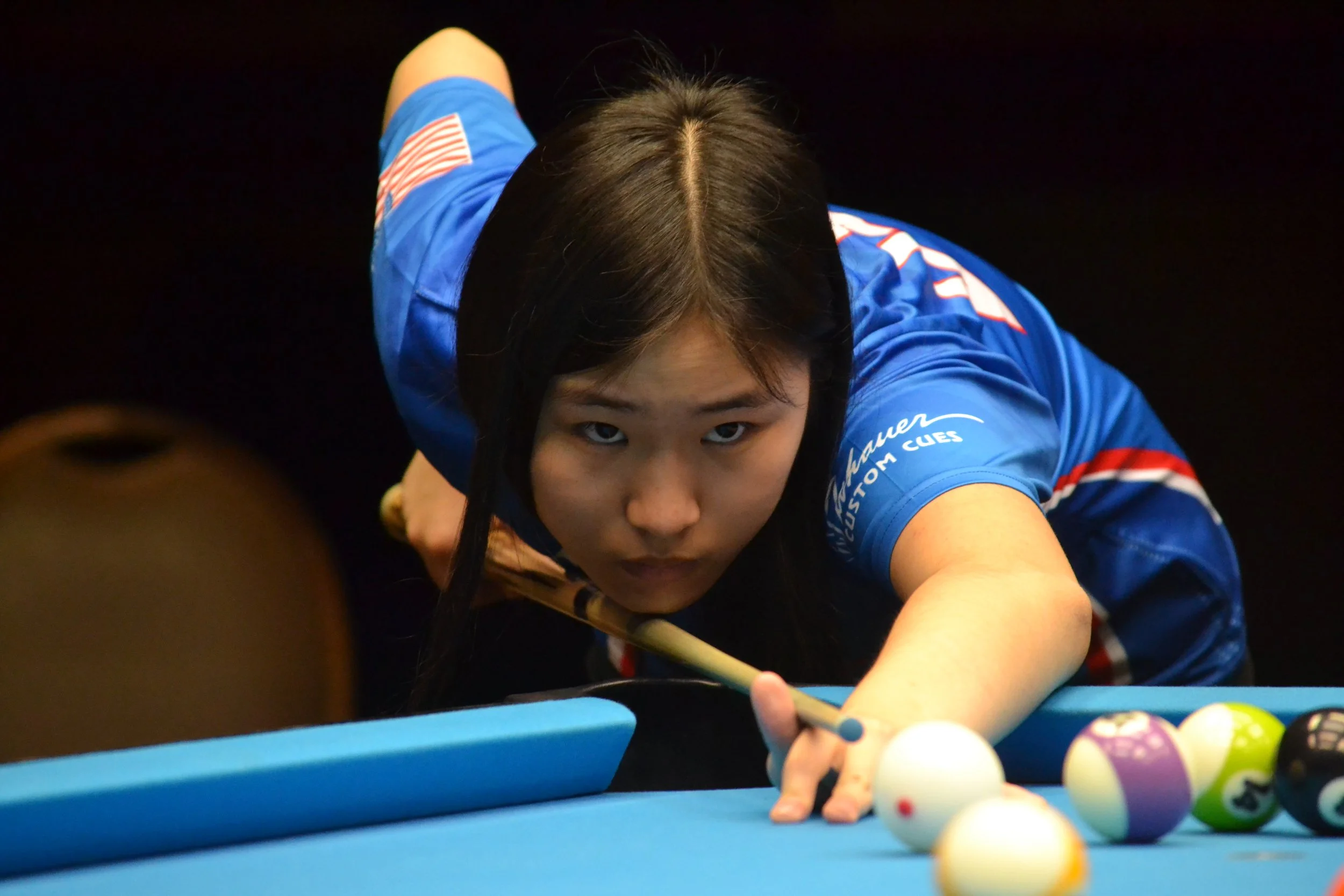 A woman playing pool, aiming to take a shot, with pool balls on the table and a focused expression.