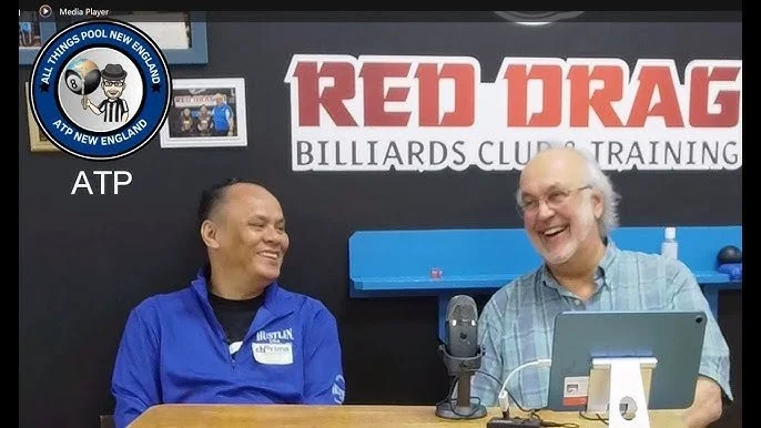 Two men smiling and sitting at a table in front of a sign that says "Red Dragon Billiards Club & Training." The man on the left is wearing a blue jacket, and the man on the right is wearing glasses and a plaid shirt. There are microphones, a laptop, and a computer monitor on the table.
