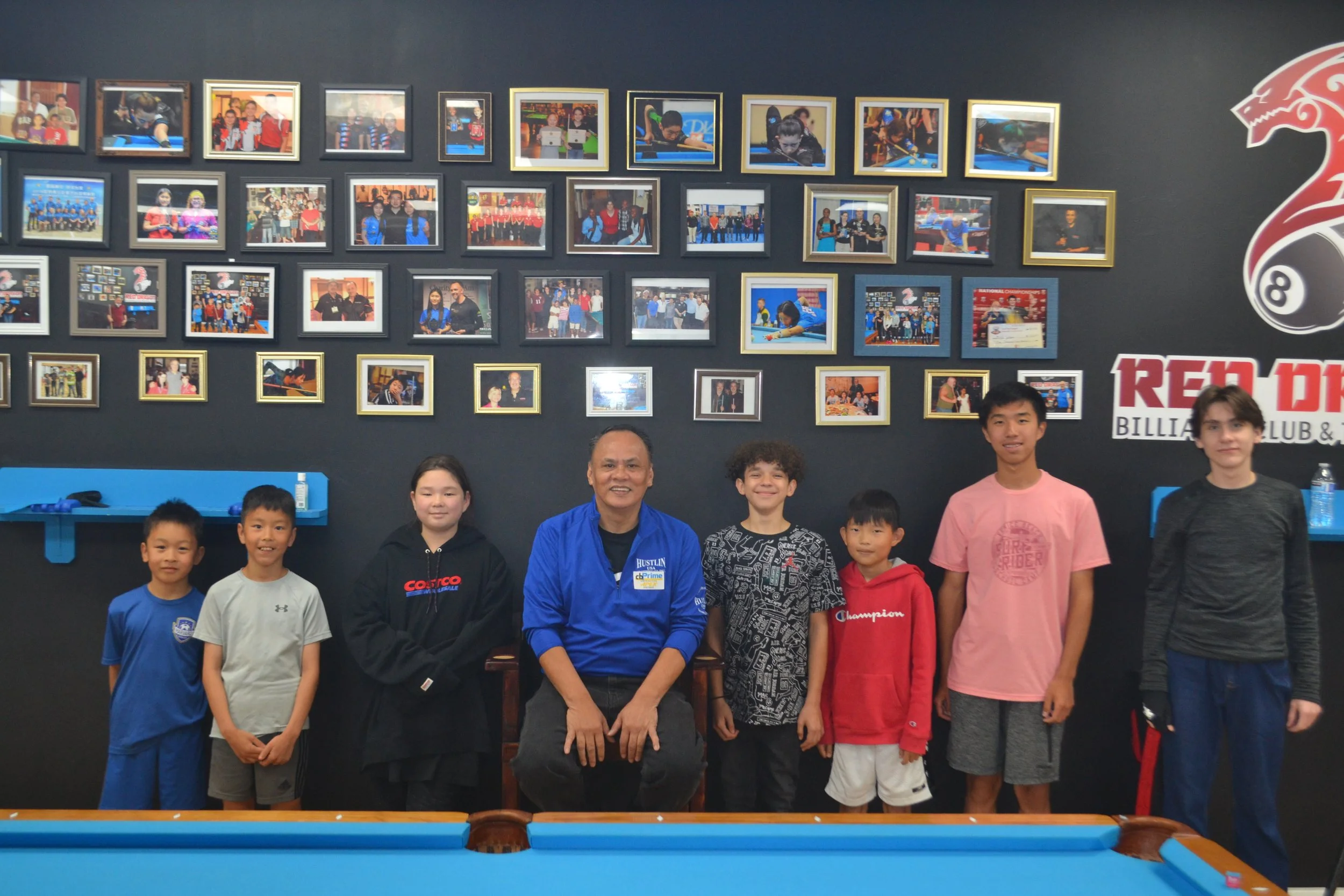 A group of seven children and two adults standing in front of a wall decorated with framed photos, billiard-themed logos, including a red dragon and an 8-ball. The setting appears to be a billiards club or activity center.