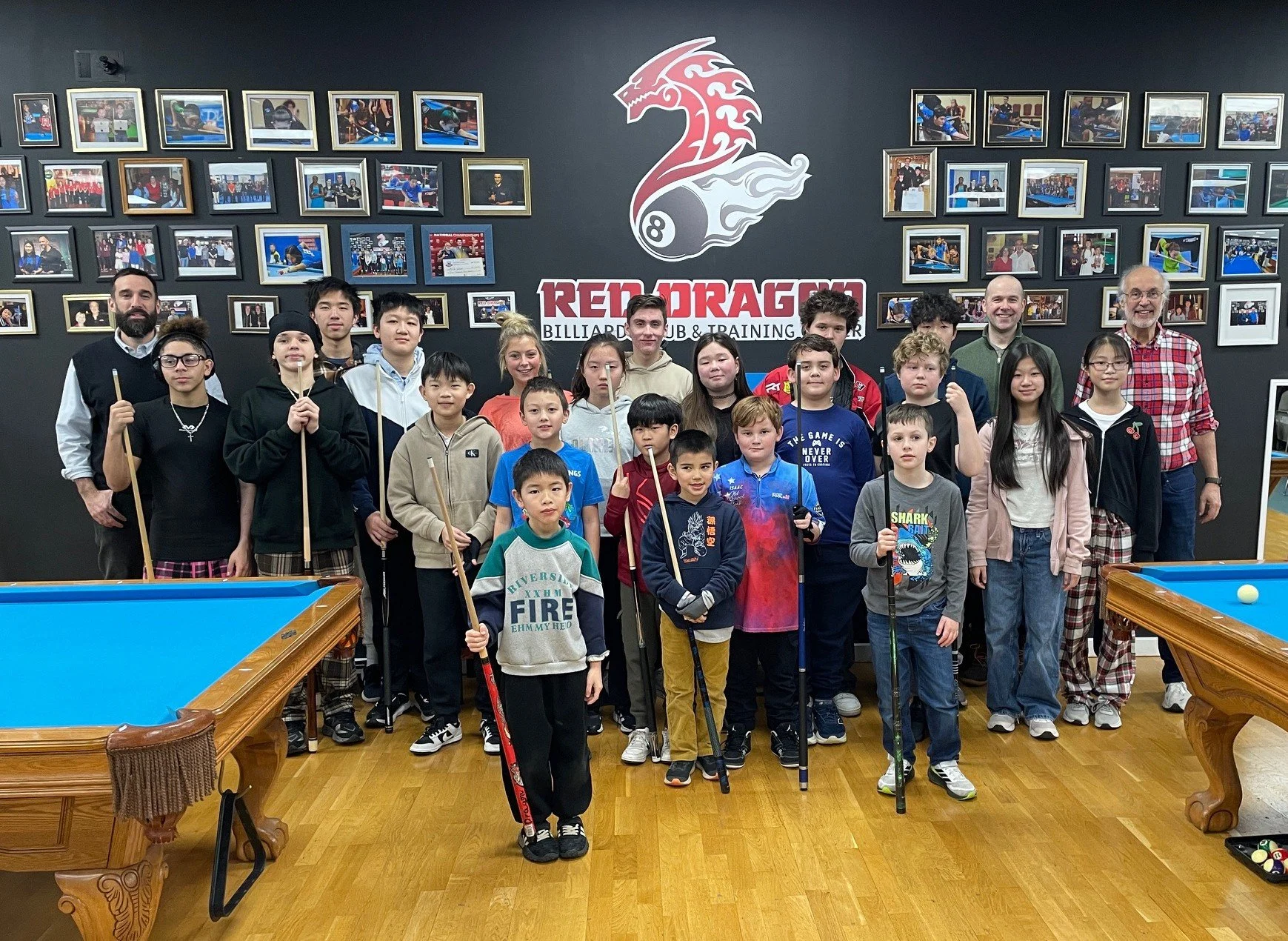 Group of children and adults standing together in a billiards club, holding pool cues, with framed photos on the wall behind them and a large logo that says 'Red Dragon' on the wall.