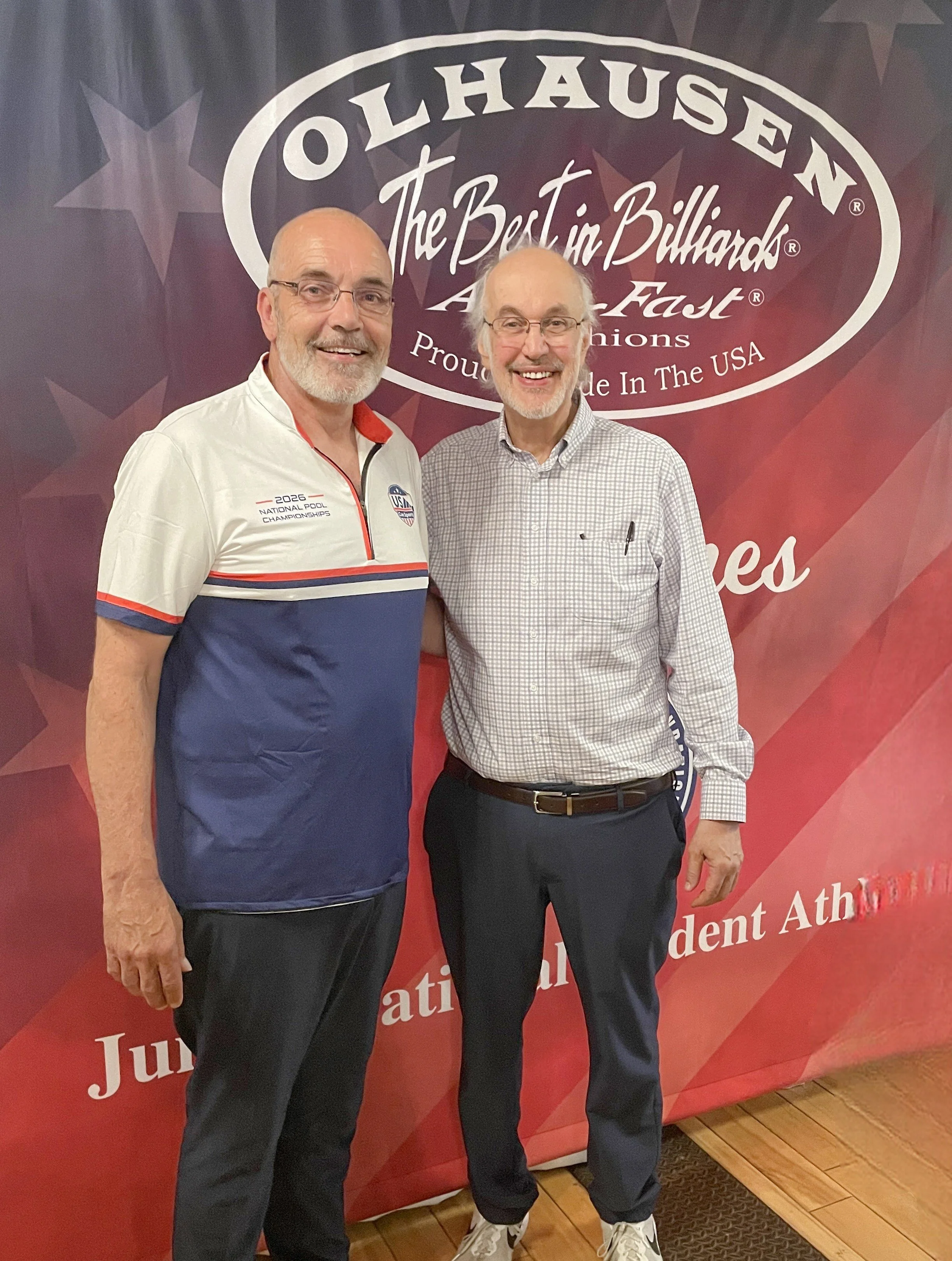 Two smiling men standing side by side in front of a red backdrop with white text and stars. One man is wearing a white polo shirt with red and blue stripes and glasses, the other is wearing a checkered shirt and glasses. They are indoors on a wooden floor.