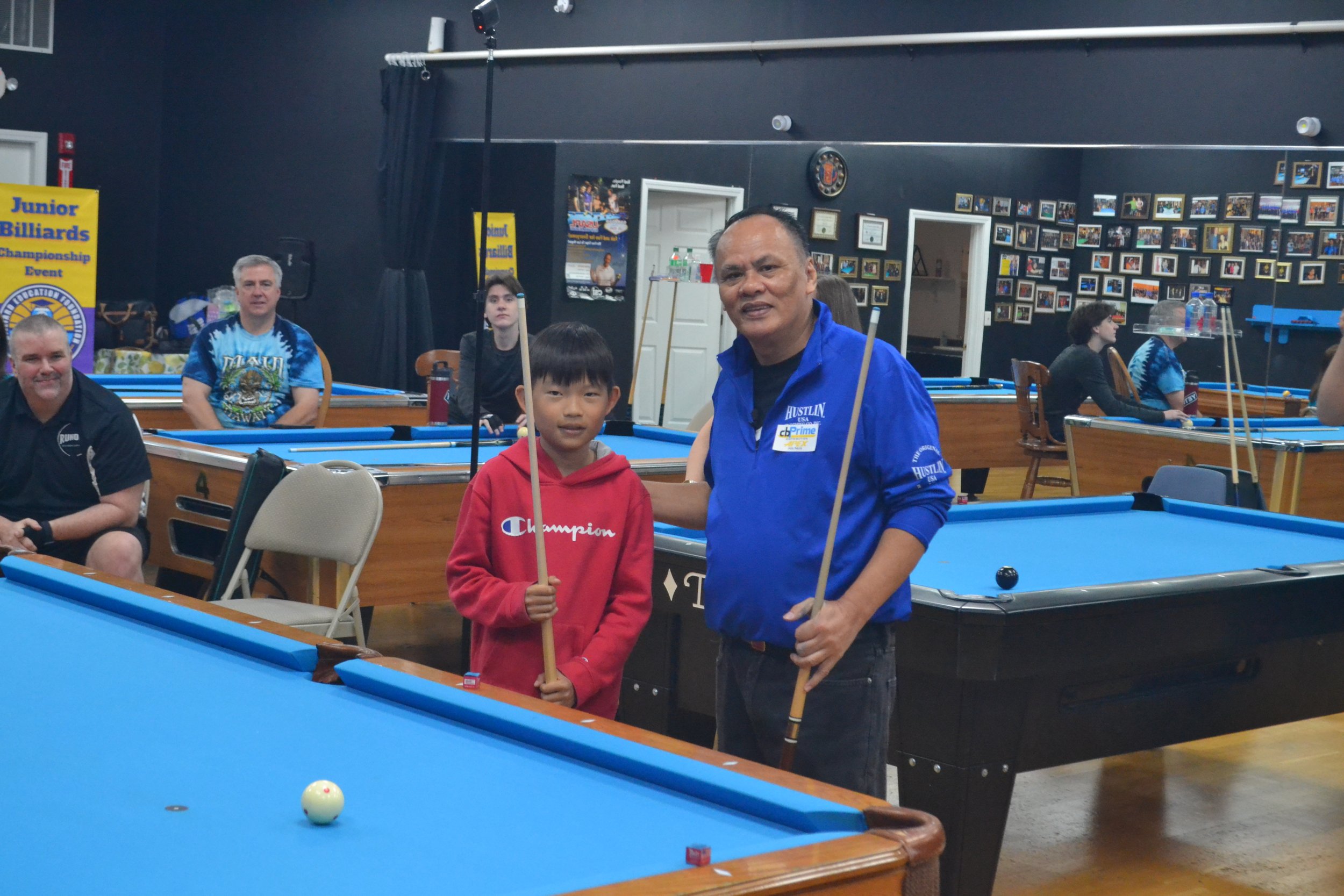 A group of people at a pool hall, with two individuals in the foreground holding pool cues, smiling for the camera. The background features pool tables, framed photos, and signs, including one for a Junior Billiards Championship Event.