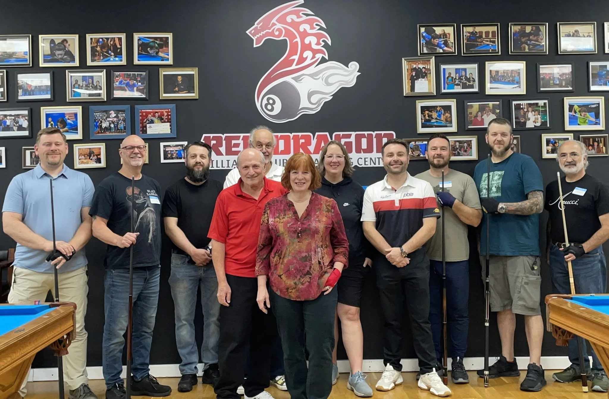 Group of people posing in a billiards hall with a large logo that reads 'Red Dragon' and photos of billiards players on the wall behind them.