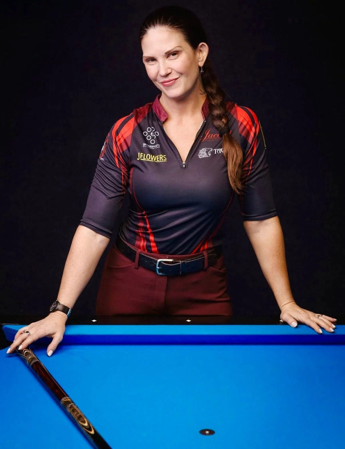 A woman standing behind a blue pool table, holding a pool cue, wearing a black and red sports shirt with logos, smiling at the camera.