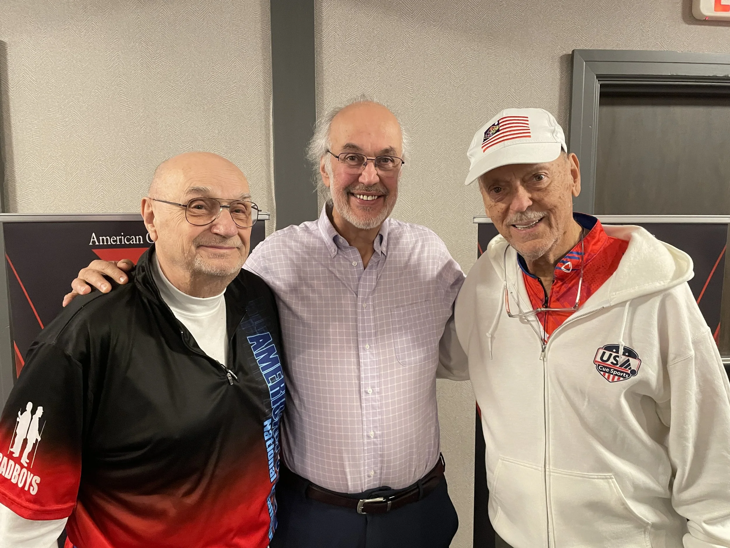 Three men smiling at the camera, standing close together indoors. Two of the men are wearing glasses, and one is dressed in a white jacket with American flags and USA Cue Sports patches. The background includes a wall with a textured pattern and a red and black banner that appears to be related to American cue sports.