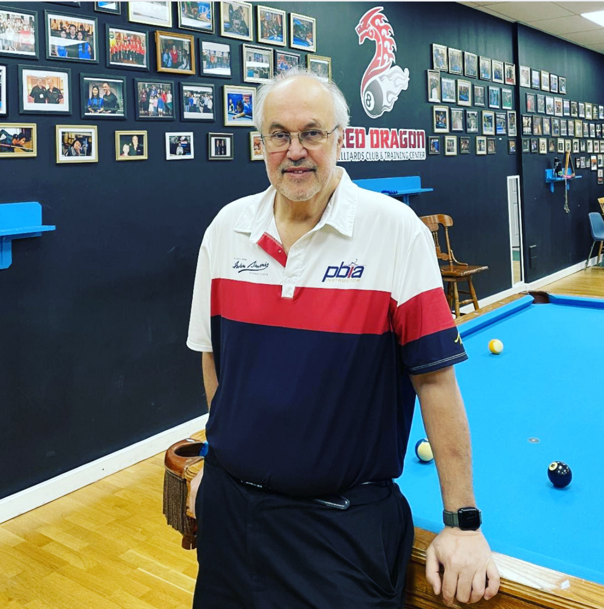 Master Instructor Roy Pastor, in a striped polo shirt standing next to a pool table in a billiards club, with walls decorated with numerous framed photographs and a logo that reads 'Red Dragon Billiards Club & Training Center.'