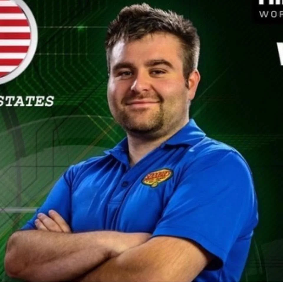 A man with short brown hair and a beard, wearing a blue collared shirt with a logo, standing with arms crossed in front of a green background with a partial American flag and text.