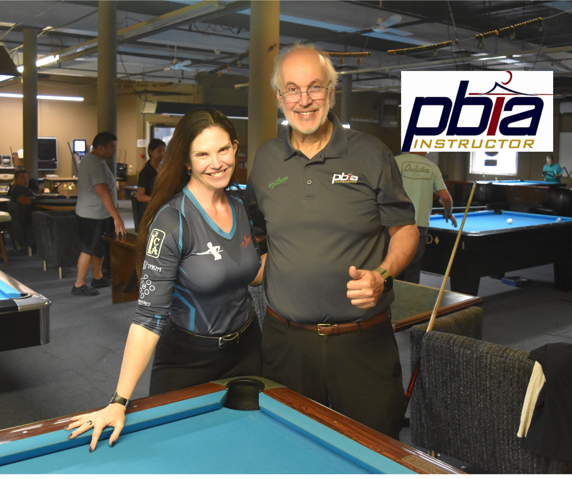 Two smiling individuals, a woman in a dark blue and black billiards jersey and a man in a gray polo shirt with a pool instructor logo, posing together at a pool hall with pool tables and players in the background, and a large logo with the text 'pba INSTRUCTOR' overlaying the image.