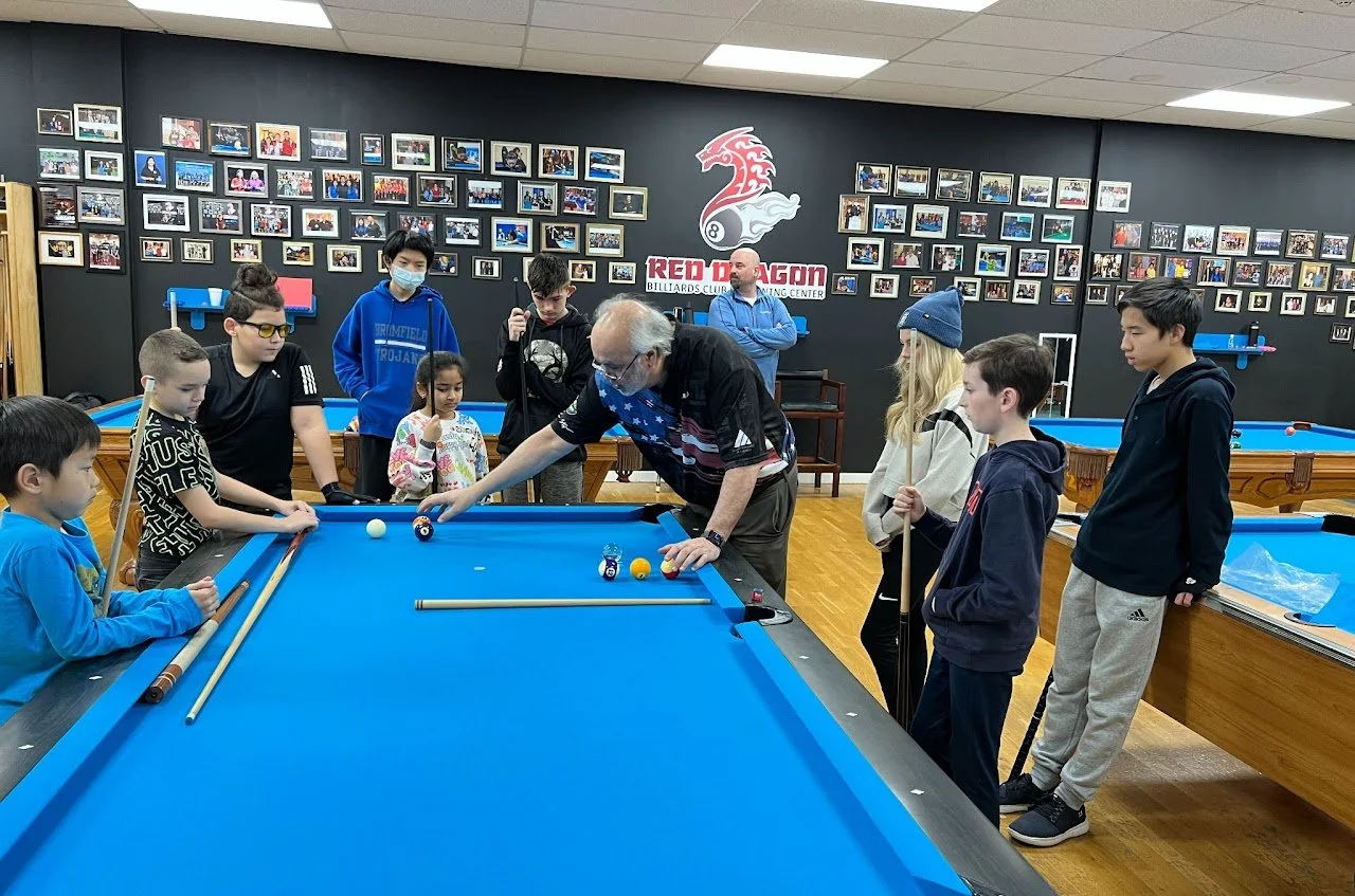 Group of children and a man playing billiards in a game room with photo collage on wall and a logo that says Red Dragon.