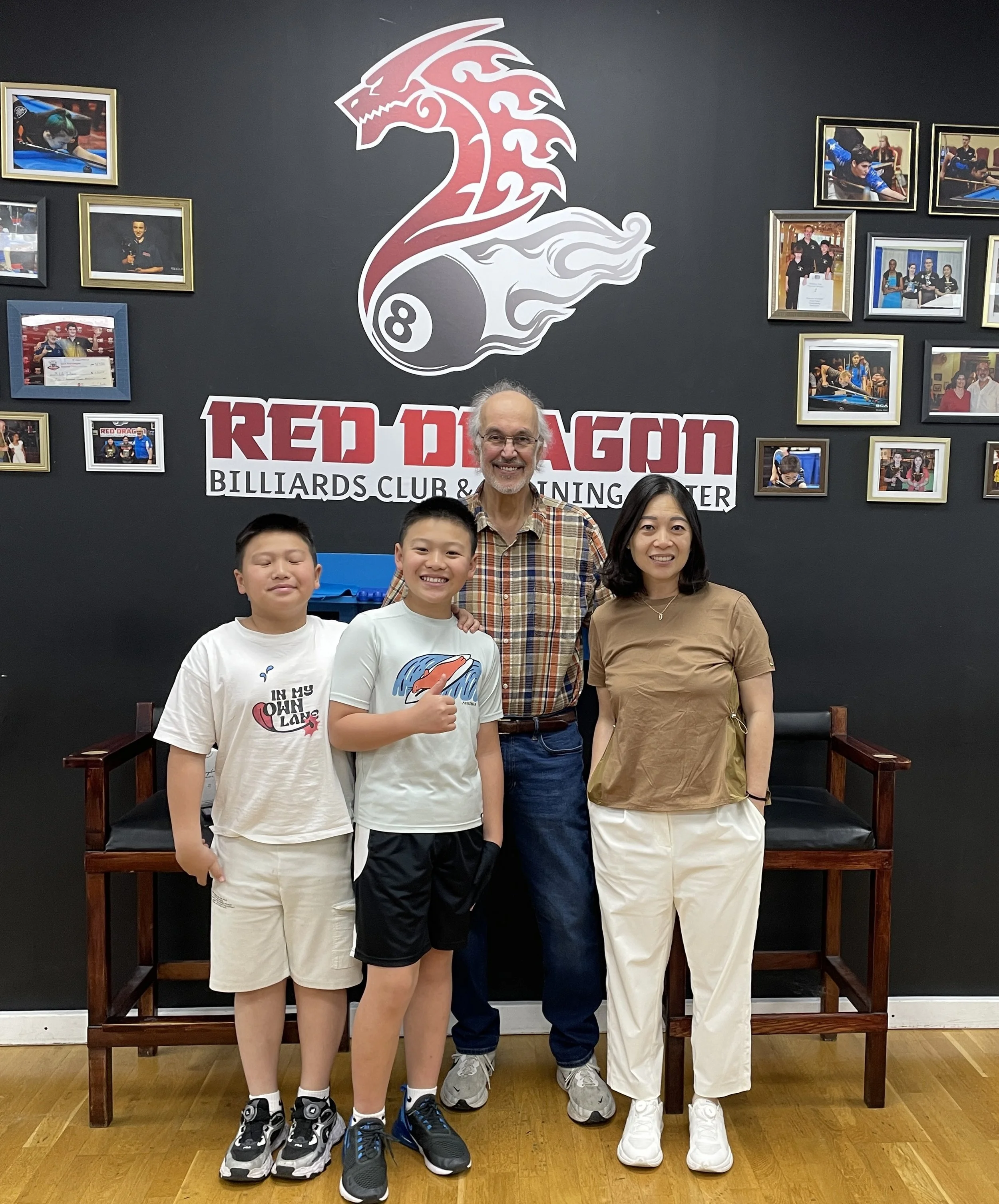 A group of five people, three children and two adults, standing in front of a wall with a red dragon logo and framed photos. The children are wearing casual clothes and sneakers, and the adults are dressed casually. They are smiling and posing for the photo.