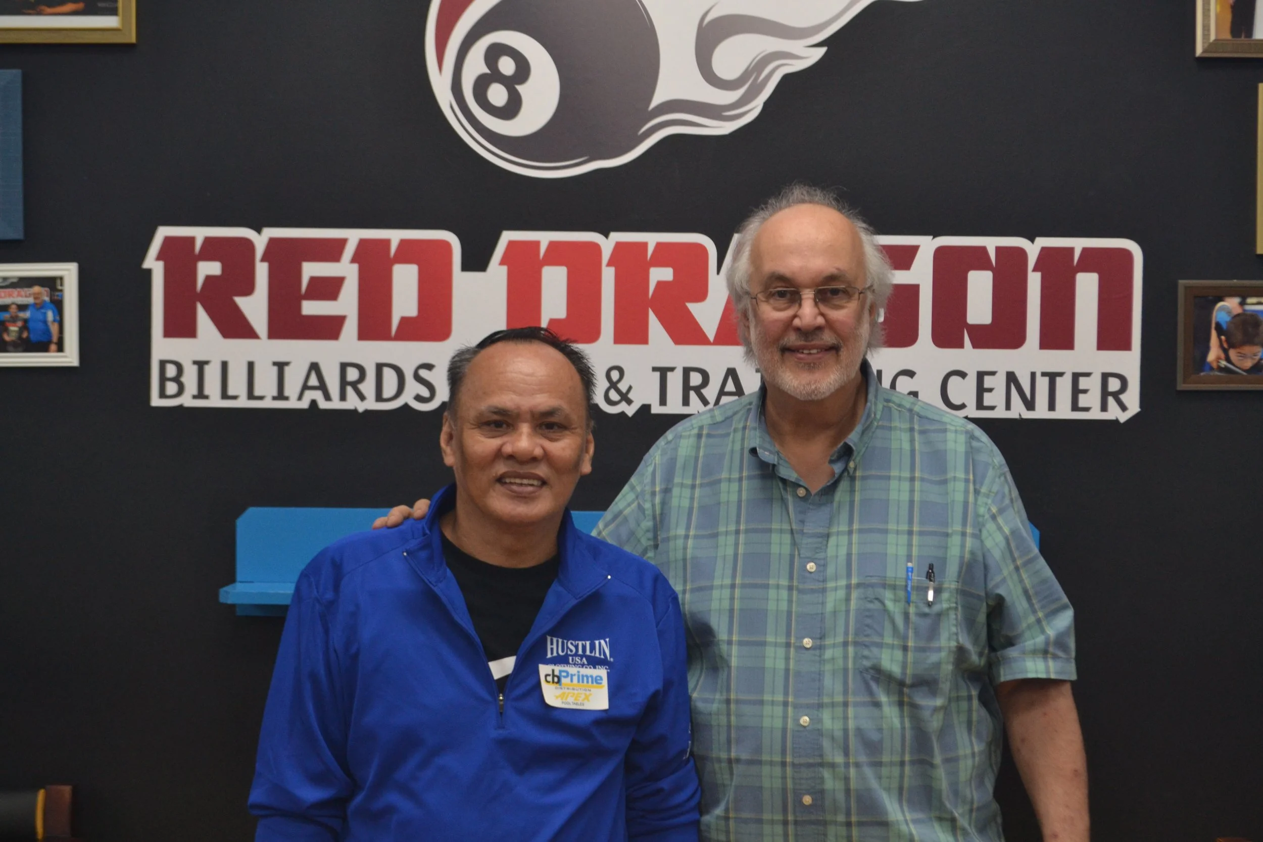 Two men smiling and standing close together in front of a sign that reads 'Red Robin Billiards & Trading Center,' with framed photographs on the wall. One man is wearing a blue jacket with a name tag, the other is wearing a green plaid shirt.