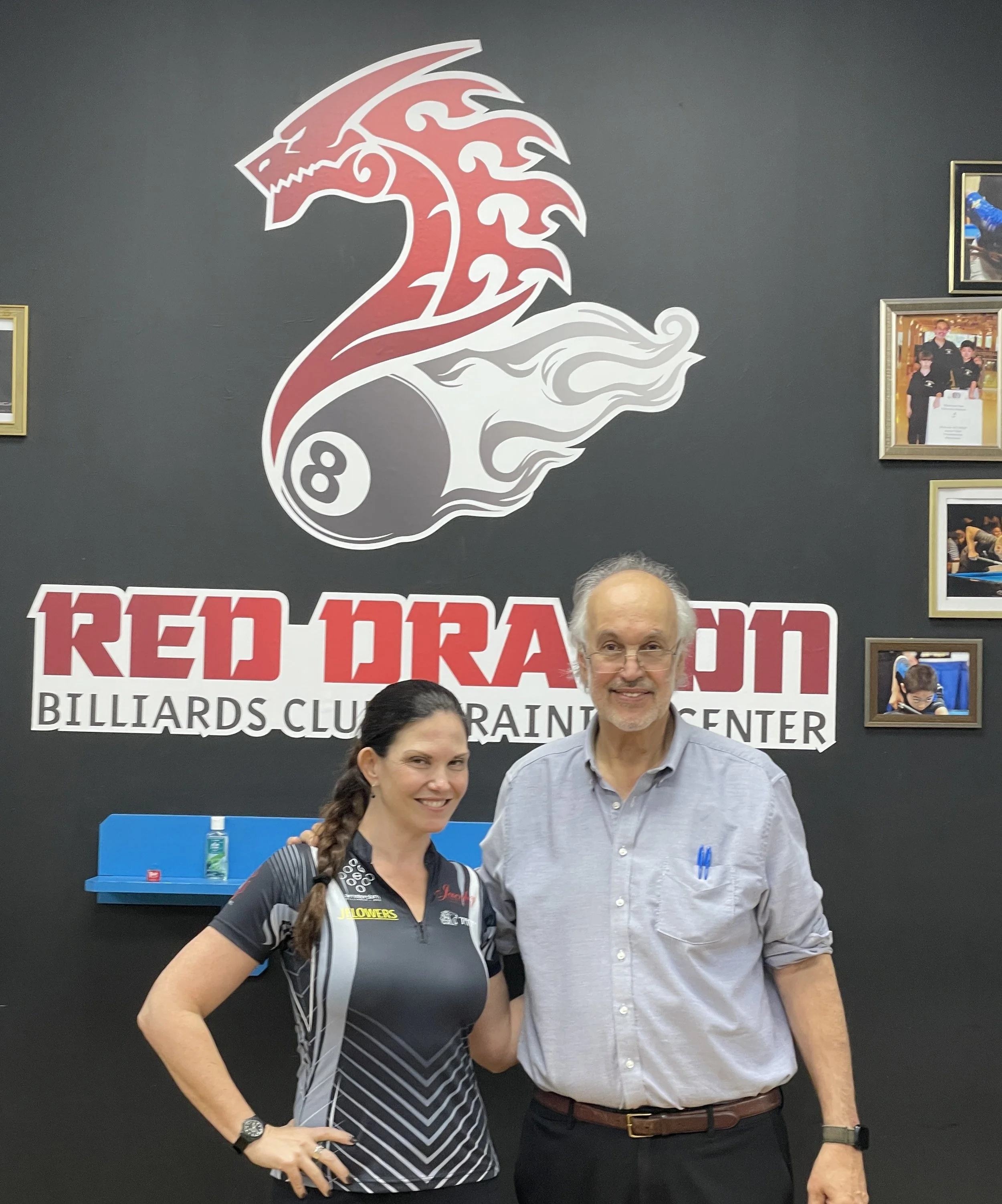 Two people, a woman and a man, stand together in front of a black wall with a large red, white, and black dragon logo and partially visible text that reads 'Red Dragon Billiards Club.' The woman has long dark hair in a braid, is wearing a black and gray sports shirt with sponsor logos, and is smiling with her hand on her hip. The man has short gray hair, glasses, and is wearing a light gray button-up shirt with pens in his shirt pocket, and is smiling.