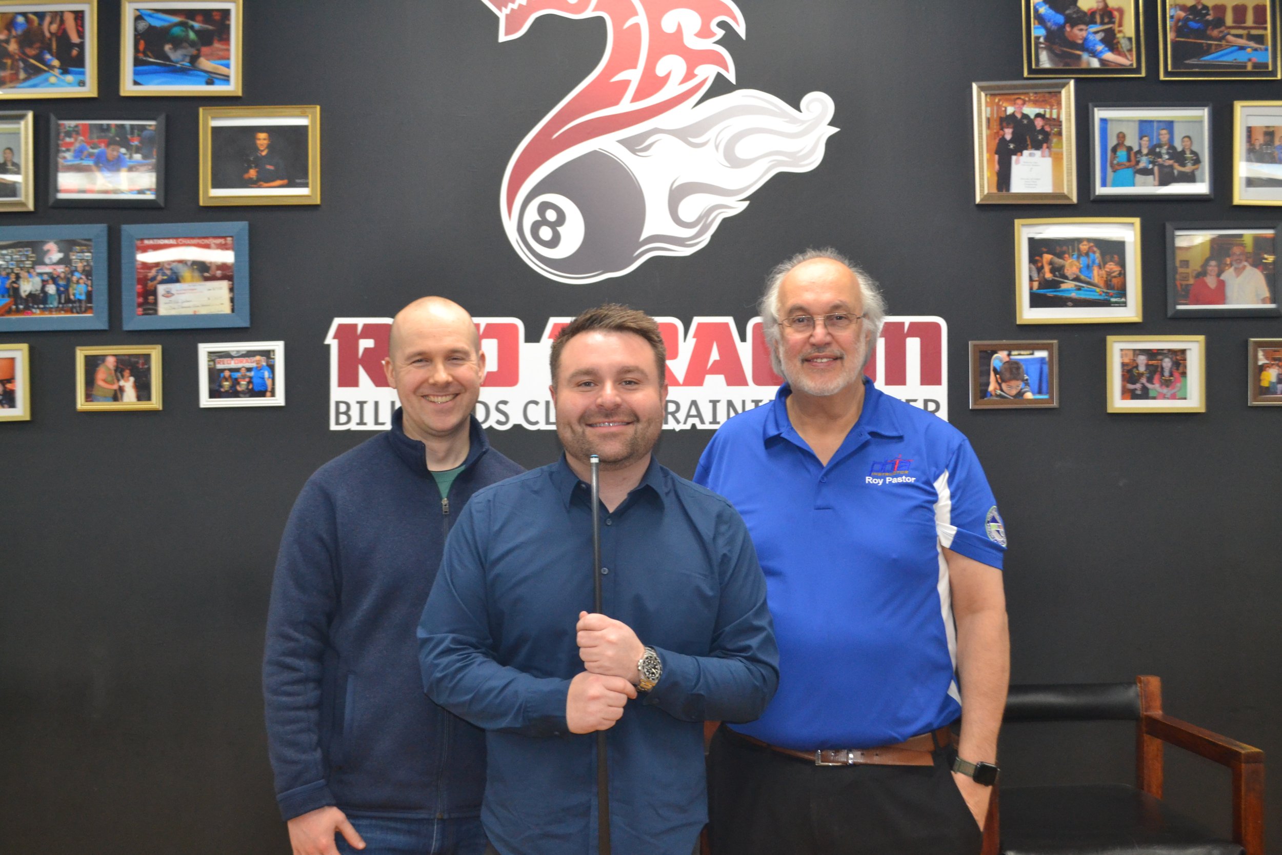 Three men standing in front of a wall decorated with framed photos of pool players. The man in the middle is holding a cue stick and smiling. The man on the right is wearing a blue polo shirt with a logo and name tag. The wall features a large logo with an 8-ball and a stylized dragon. The scene appears to be at a billiards club or tournament.