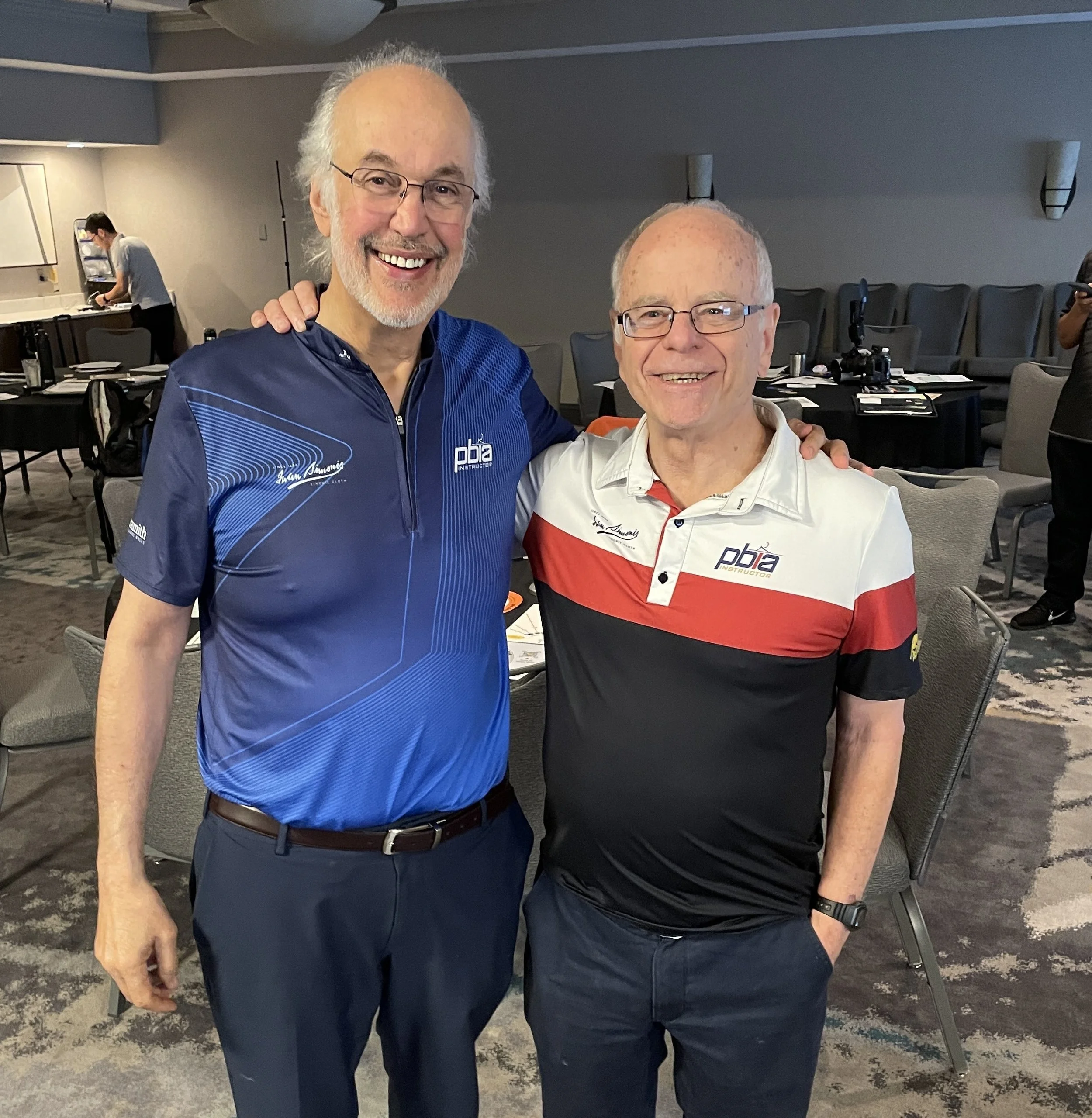 Two smiling men in casual clothing standing close together in a conference room, with empty chairs and tables in the background. One man has white hair and glasses, the other has short hair and is also wearing glasses.