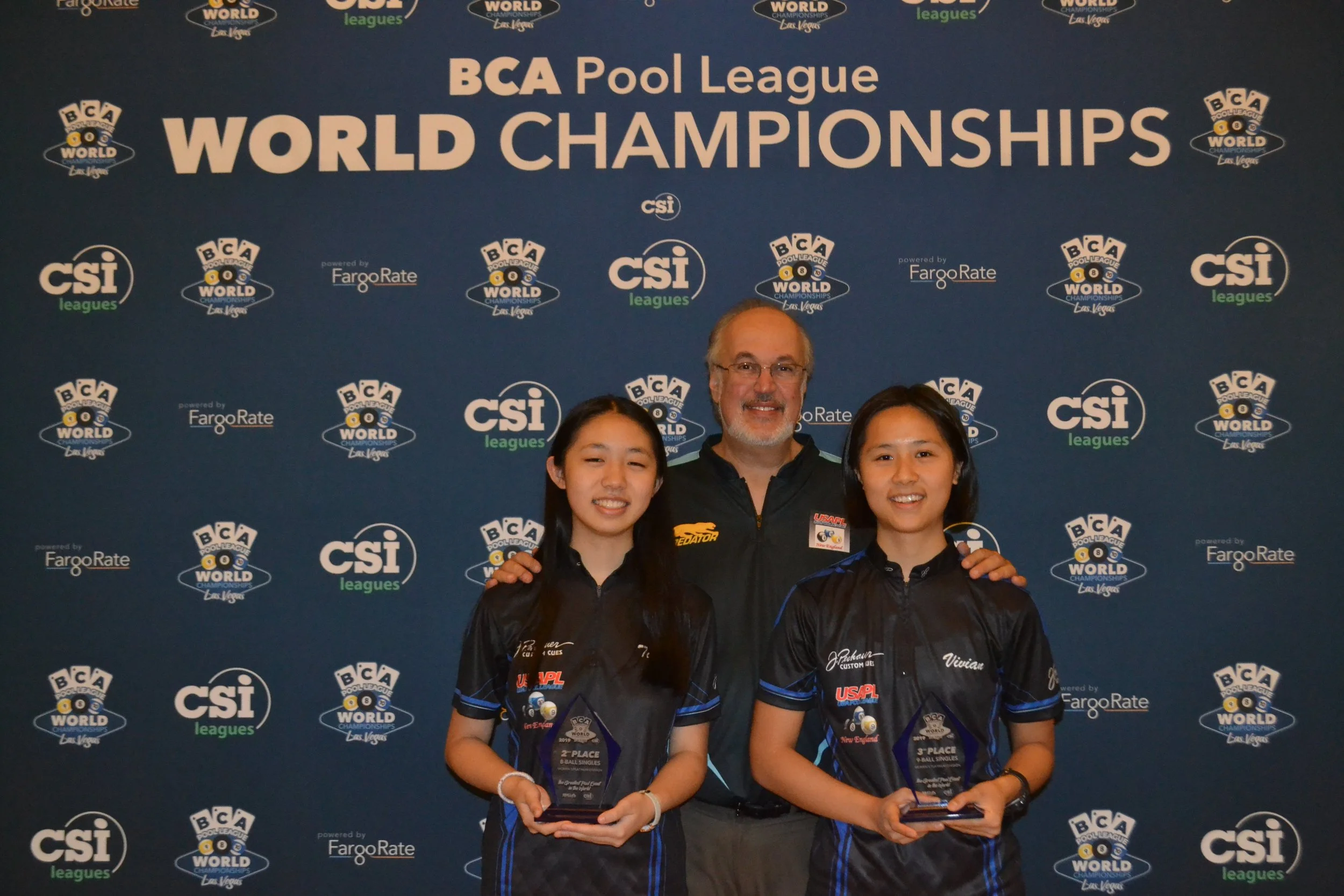 Three people, two young women and one older man, smiling and posing with awards in front of a backdrop with BCA Pool League and CSI logos, at the BCA Pool League World Championships in Las Vegas.