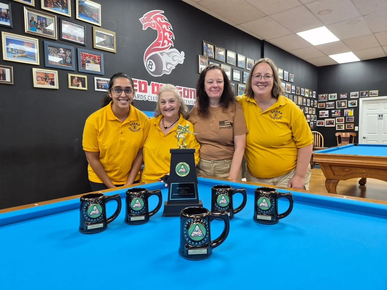 Four women posing behind a blue pool table at a billiards club, with a trophy and four black mugs with green and blue logos. The women are smiling, with the two on the left wearing yellow shirts, and the woman second from the right wearing a tan shirt. The background features framed photos and a sign that reads 'Red Dragon Billiards Club.'