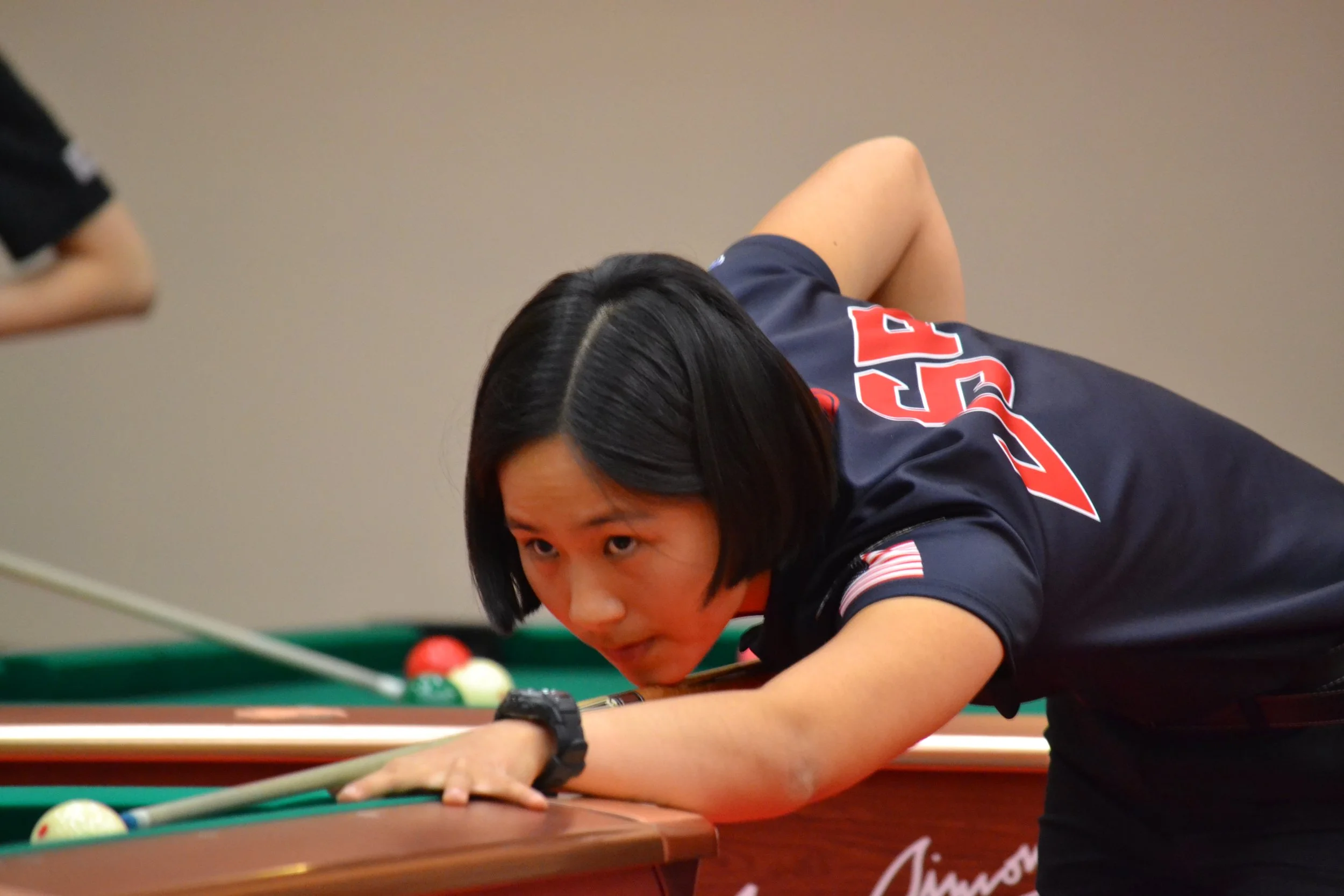 A woman with black hair, wearing a dark sports uniform, playing pool and focusing on the shot.