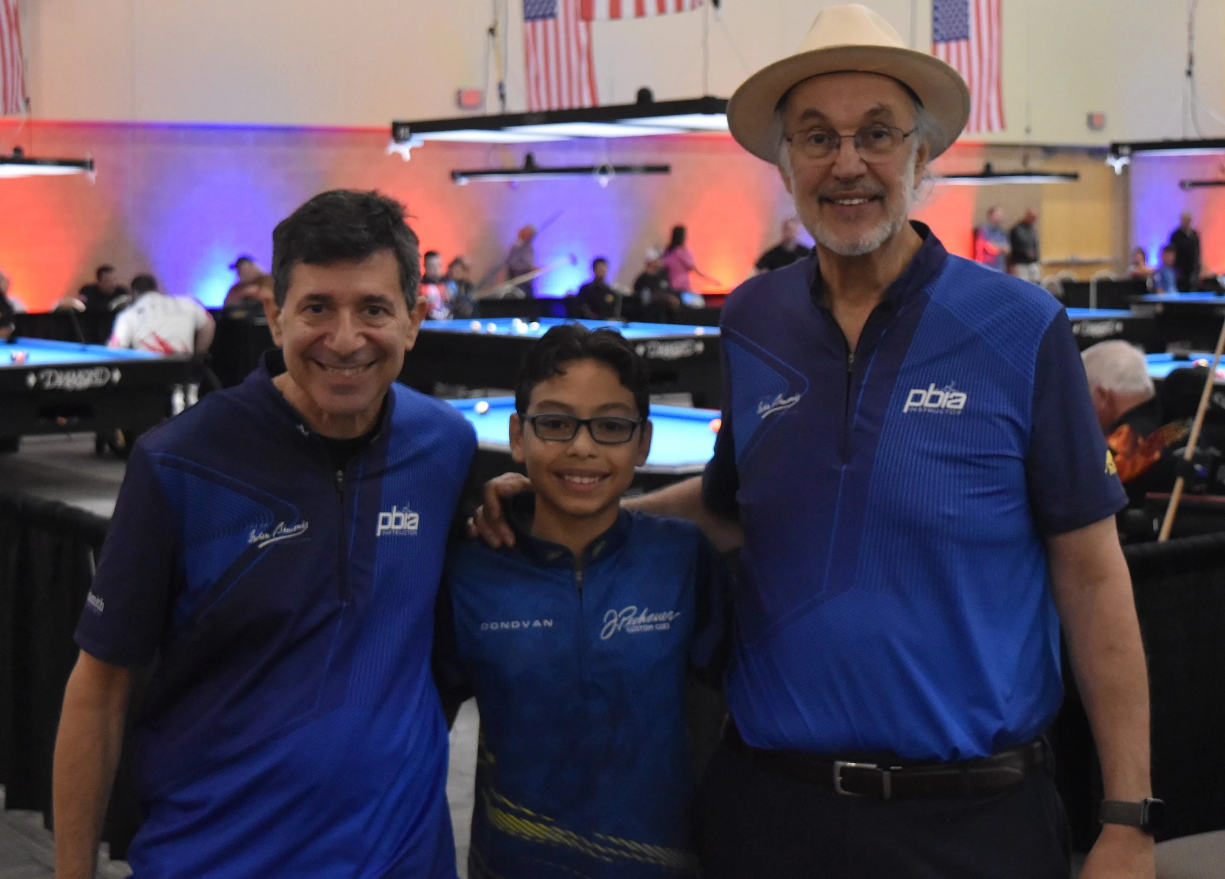 Three people in blue shirts at a pool hall with pool tables and American flags on the wall in the background.