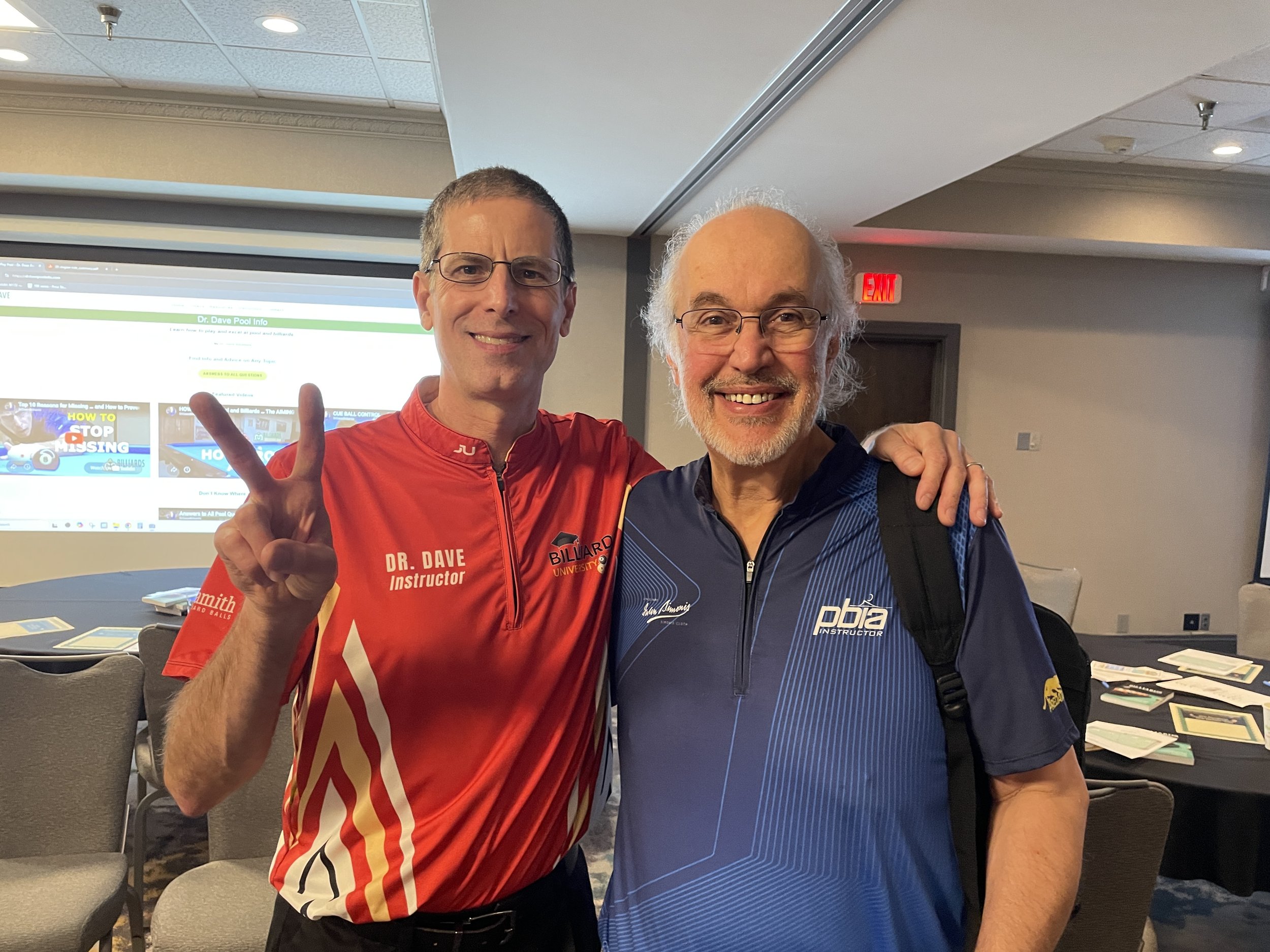 Two men smiling and posing for a photo indoors. The man on the left is wearing a red sports shirt with the name 'Dr. Dave' and 'instructor' written on it, and making a peace sign. The man on the right is wearing glasses and a dark blue shirt with text on it. Behind them are chairs and a large screen displaying a website.