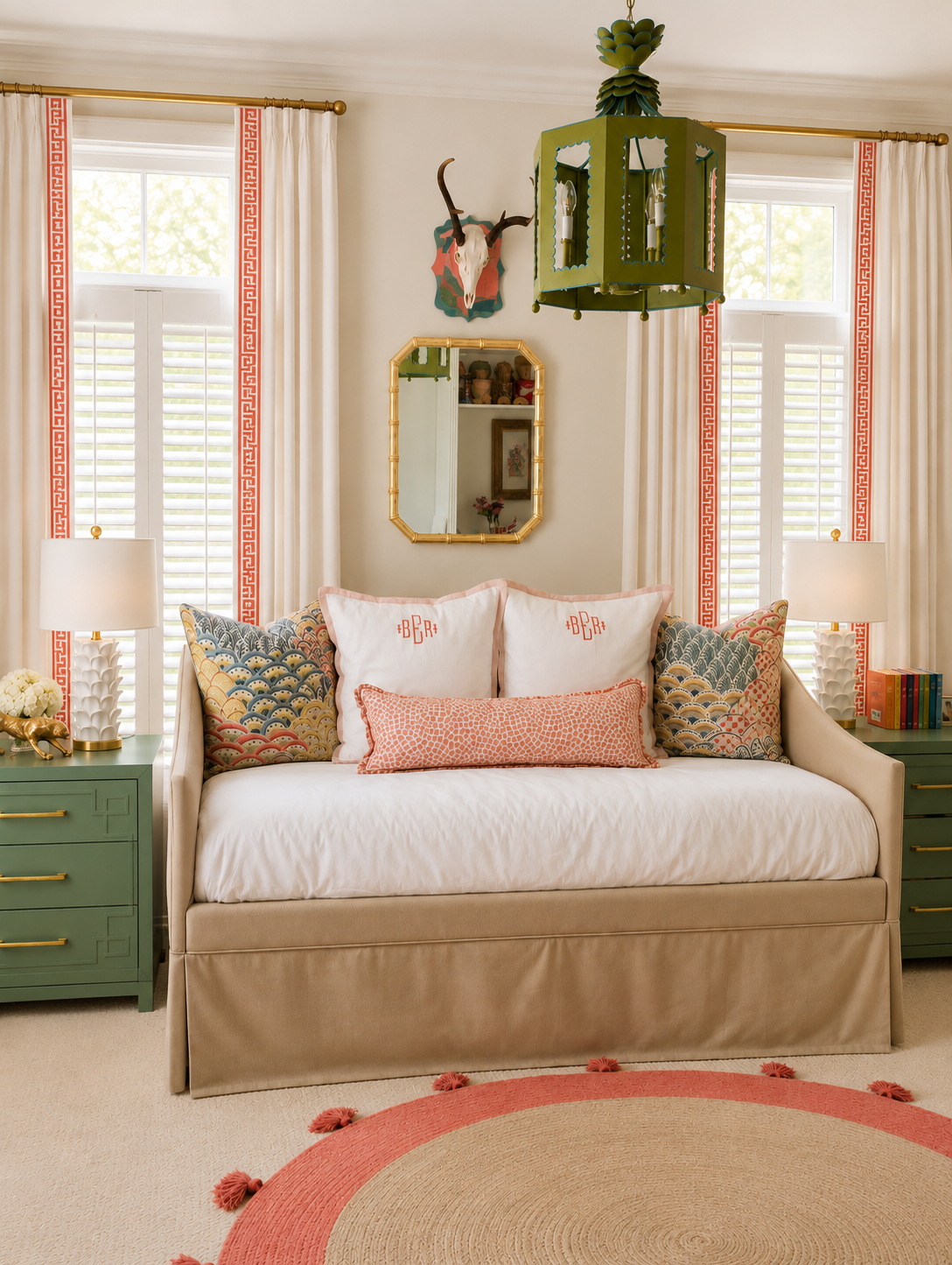 A bedroom with a bed featuring decorative pillows and a white upholstered skirt, green nightstands with lamps, a round coral-colored rug with tassels, white curtains with coral embroidery, a mirror, a mounted animal skull with horns, and a hanging green lantern-style light fixture.