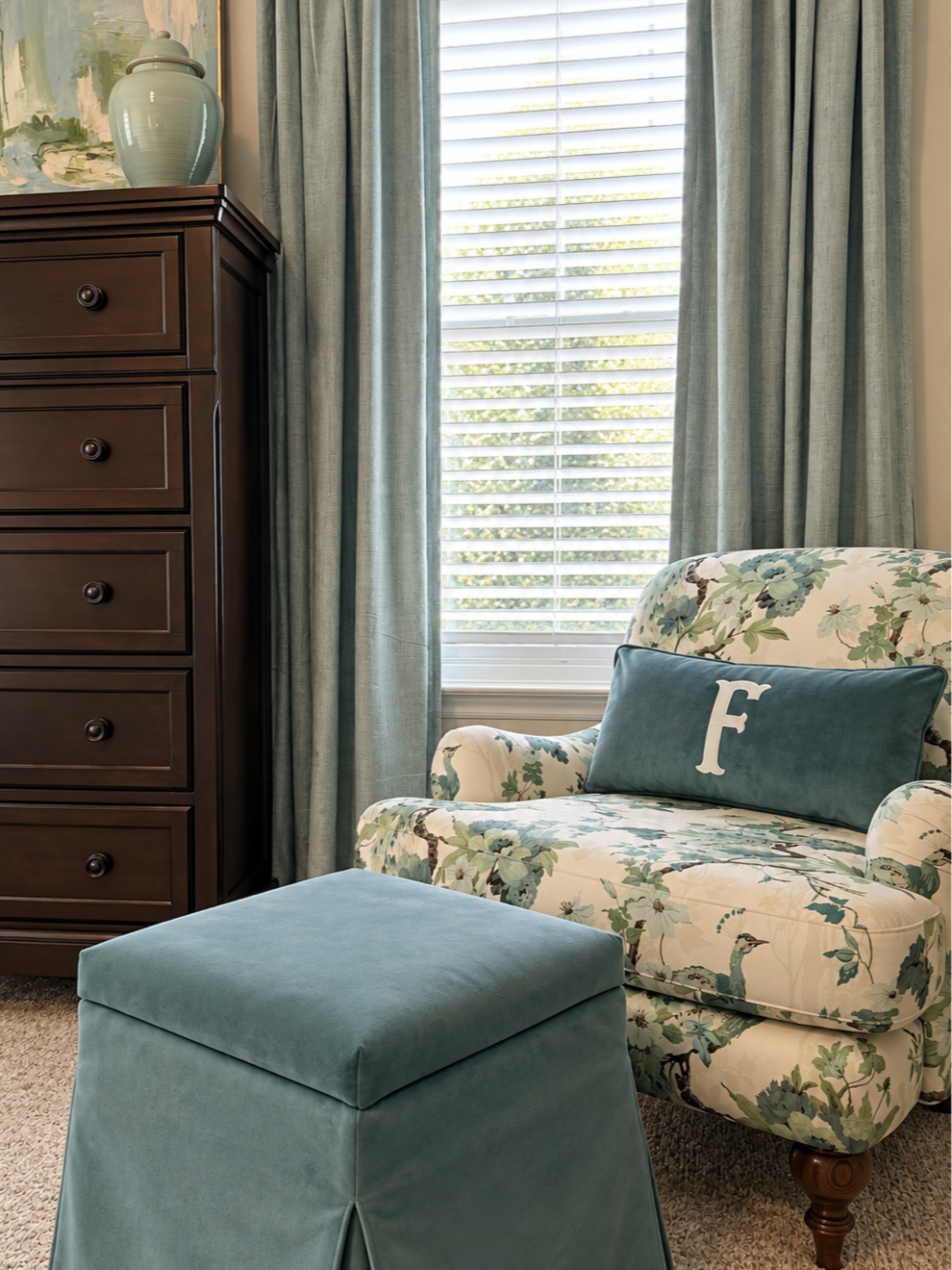 A cozy corner with a floral upholstered armchair with a blue embroidered pillow, a matching blue ottoman, a dark wooden dresser, and a window with light blue curtains and white blinds.