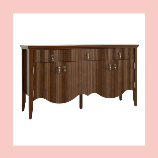 A vintage wooden sideboard with vertical grooves, brass handles, and curved bottom edge, standing against a pink background.