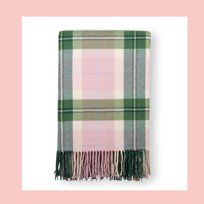 A folded plaid blanket with green, pink, and white colors, featuring fringe edges at the bottom, placed on a pink and white background.