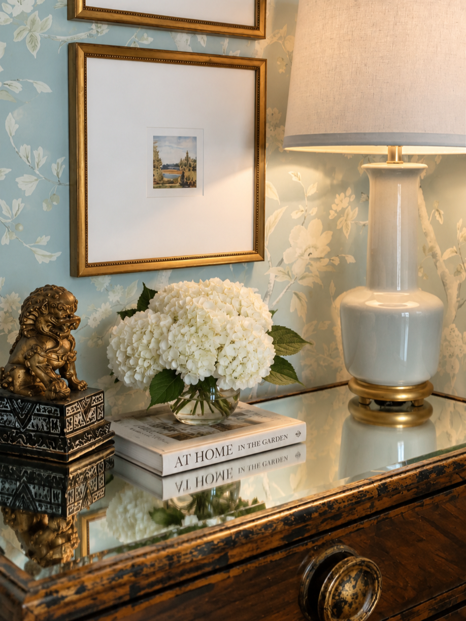Decorative table with white hydrangeas, books, a lamp, and a golden lion figurine against a floral wallpaper background.