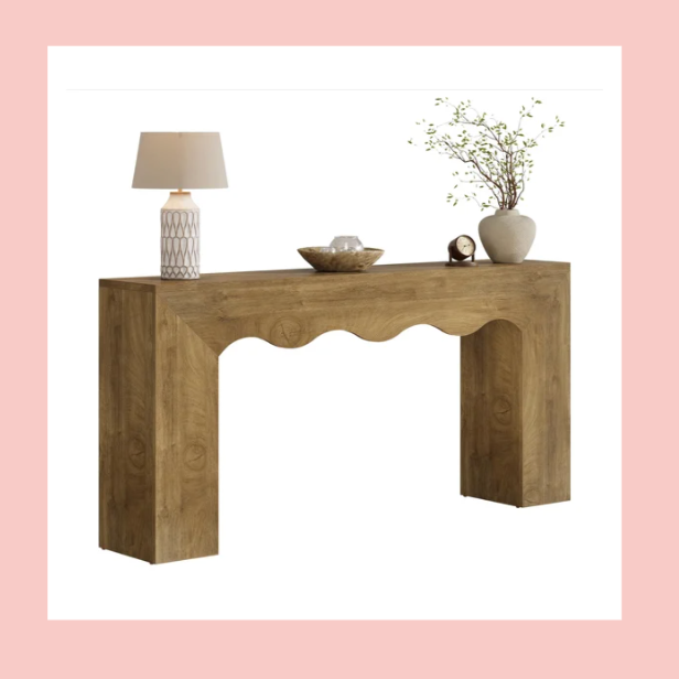 A wooden console table decorated with a table lamp, a bowl, a small clock, and a potted plant with thin branches on top.