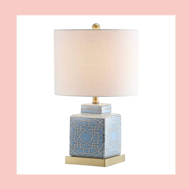Decorative table lamp with a light-colored shade and a geometric patterned base in blue and gold, placed on a gold rectangular platform.