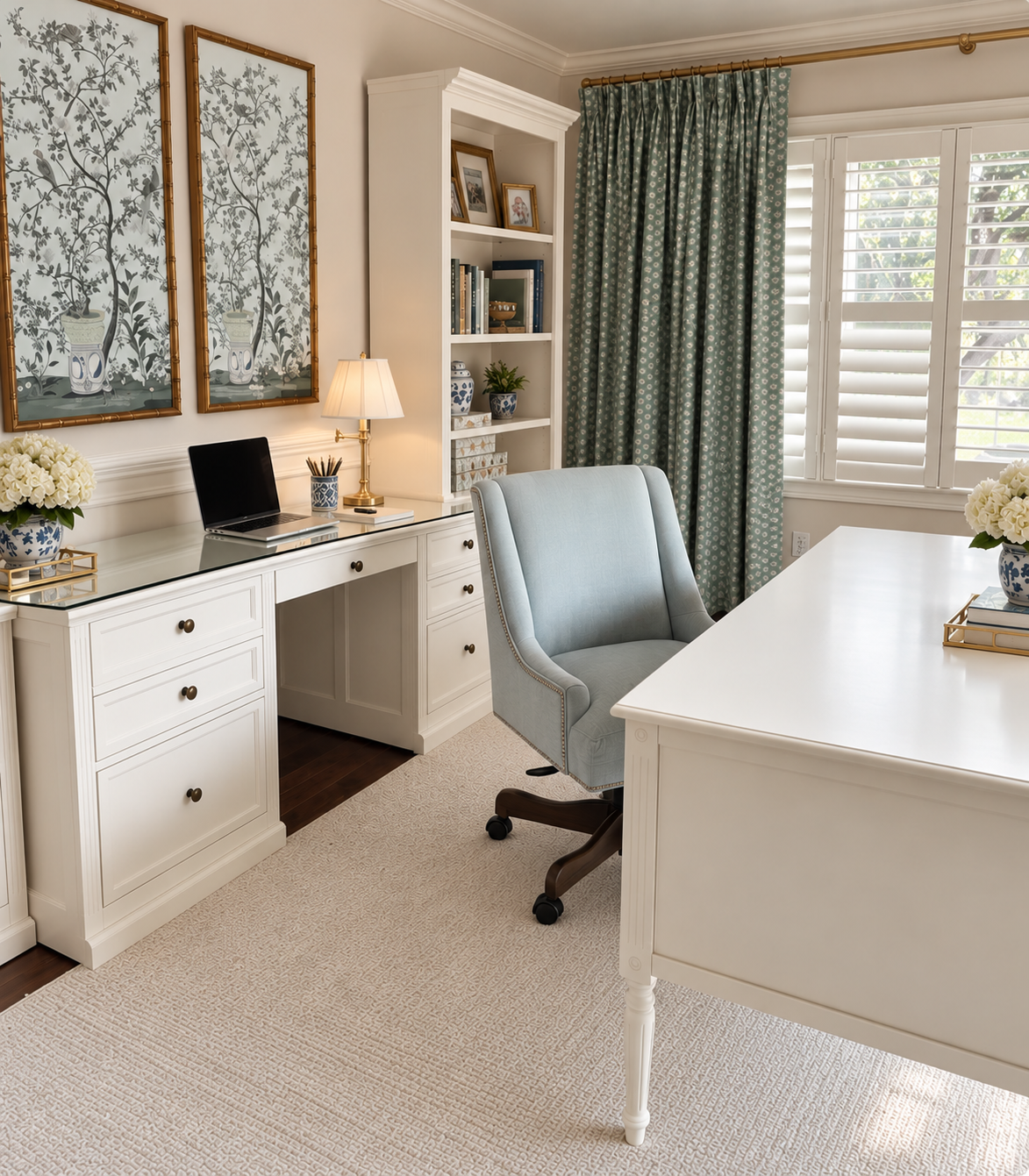 Bright and organized home office with white built-in desk, light blue swivel chair, decorative vases with white flowers, a table lamp, a bookshelf, and windows with plantation shutters and floral curtains.