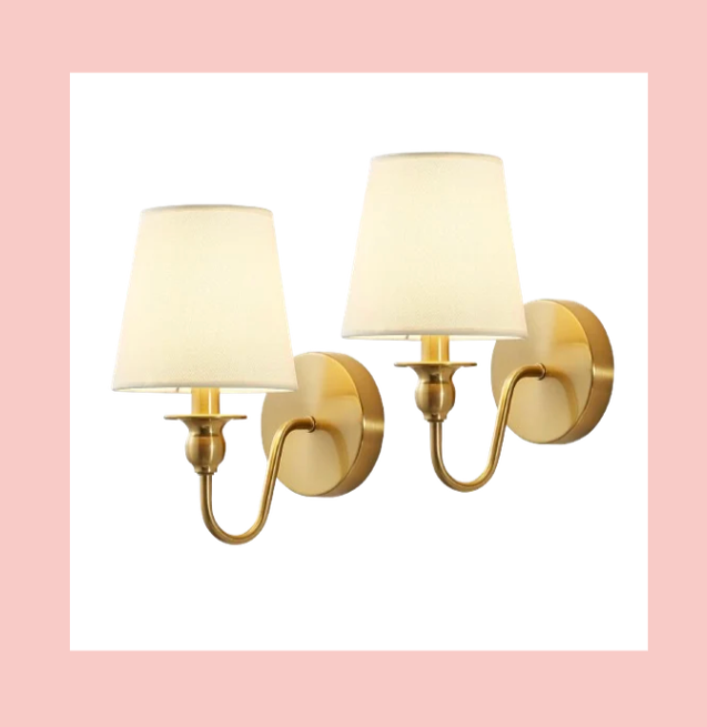 Two gold wall-mounted lamps with white lampshades on a pink background.