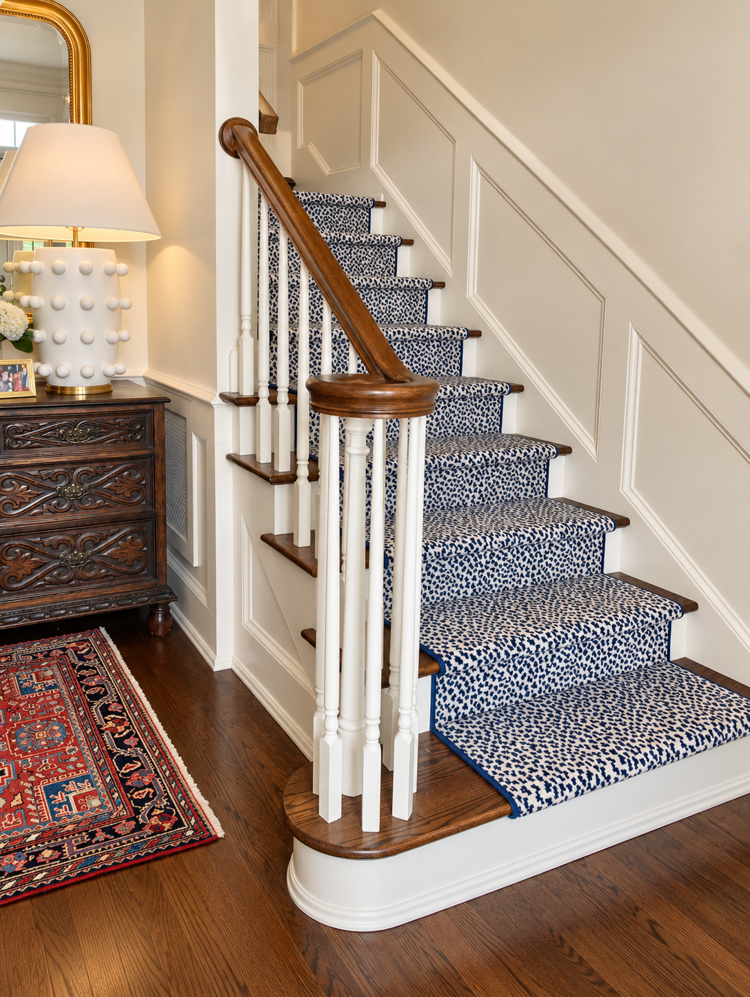 Indoor staircase with a blue and white patterned carpet, wooden handrail, and white spindles, next to a dark wooden chest of drawers with decorative carvings and a lamp with a white shade.