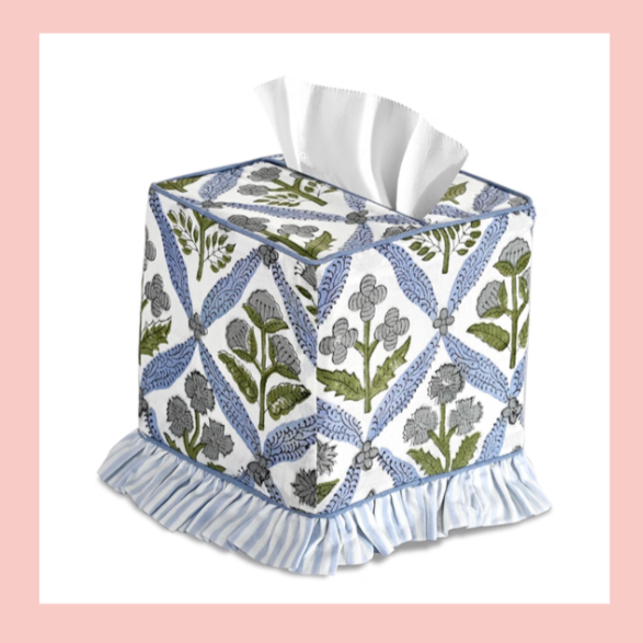 Decorative tissue box cover with a floral and leaf pattern, ruffled skirt at the bottom, and a tissue pulled out on top, set against a pink background.