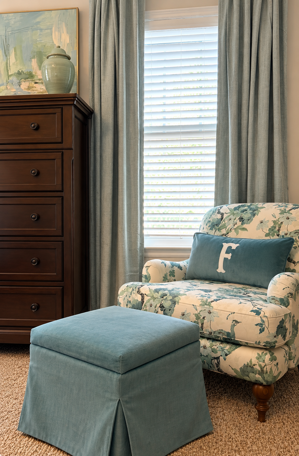 A cozy living room corner with a floral patterned armchair, a blue pillow with the letter 'F', a matching blue ottoman, a dark wood dresser, a large abstract painting, blue curtains, and window blinds letting in natural light.