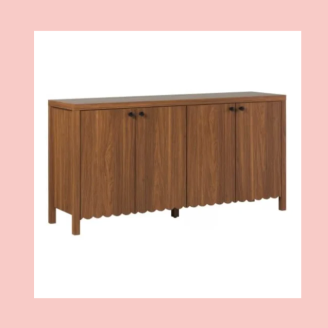 A wooden sideboard with four cabinet doors, black knobs, and a scalloped bottom edge, set against a pink and white background.