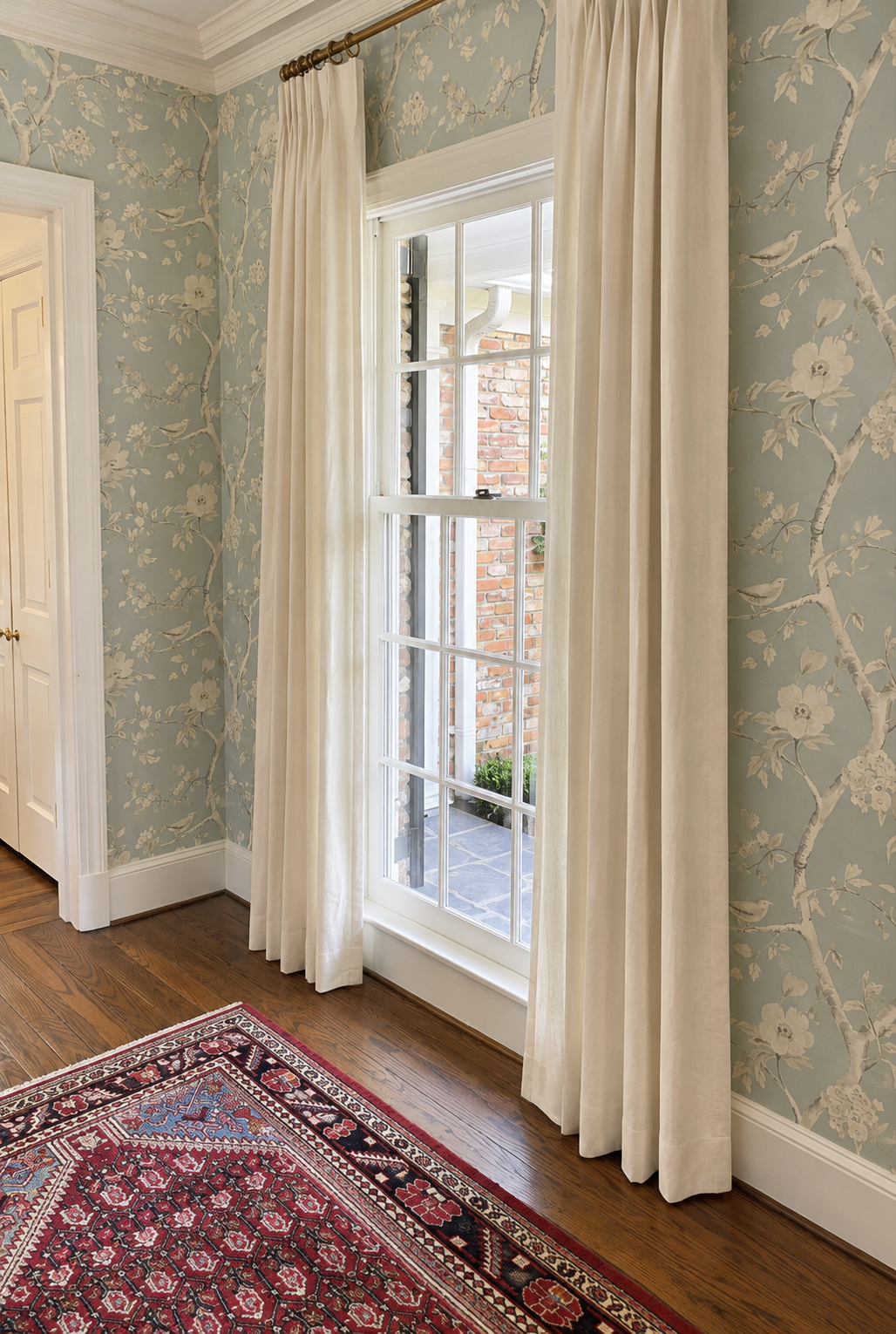 A room with floral wallpaper, a window with white curtains, a hardwood floor, and a red patterned rug.