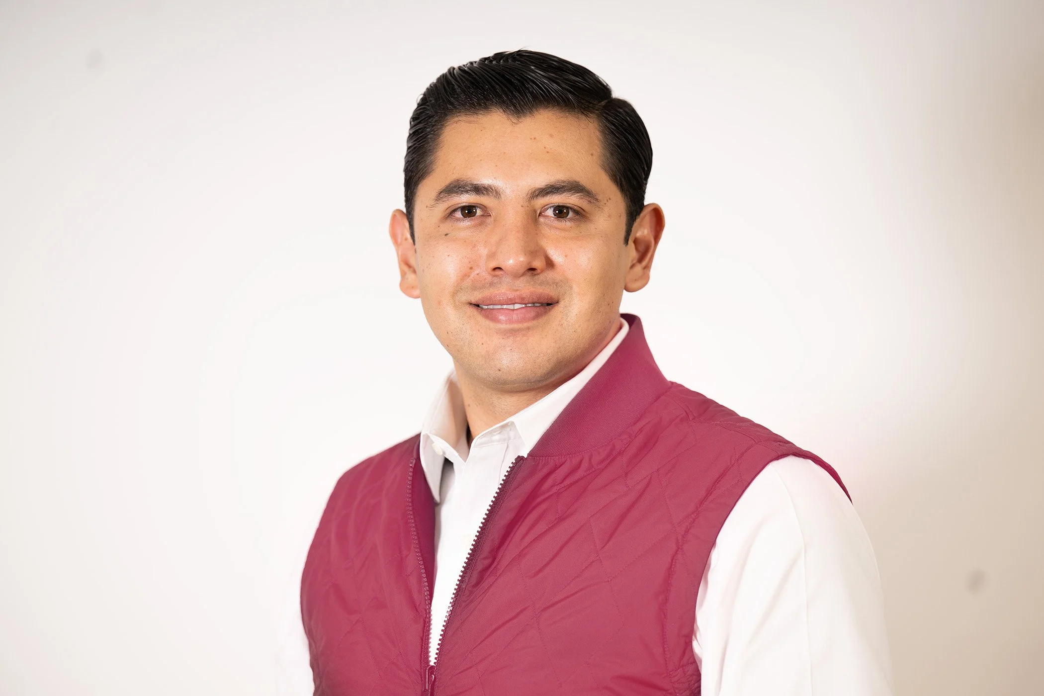 A young man with dark hair slicked back, wearing a white shirt and a maroon quilted vest, smiling softly against a plain white background.