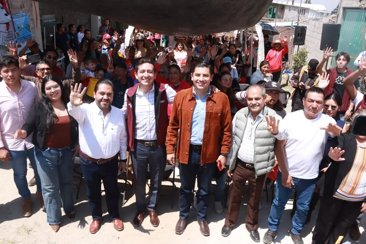 Group of people gathered outdoors, waving and smiling, with some seated and some standing under a tent.