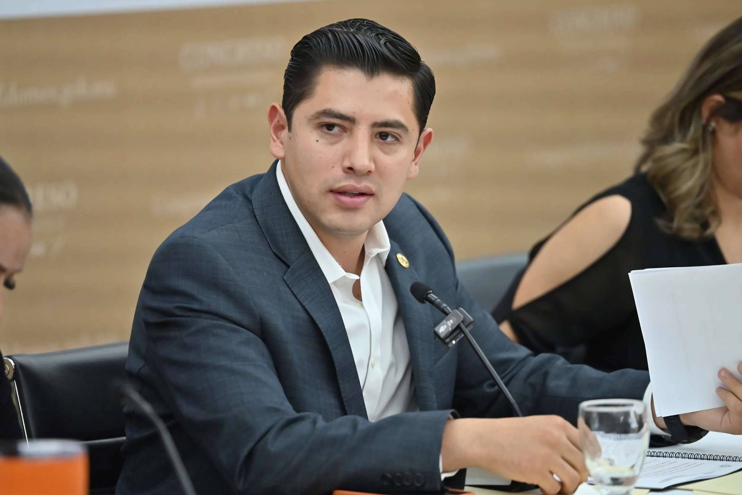 A man in a dark blue suit and white shirt seated at a table during a formal meeting or hearing. He has dark, slicked-back hair and is looking to his left. There is a microphone in front of him and papers on the table. To his right, part of a woman in black with shoulder-length hair is visible, holding papers. The background contains a blurred wooden panel wall.