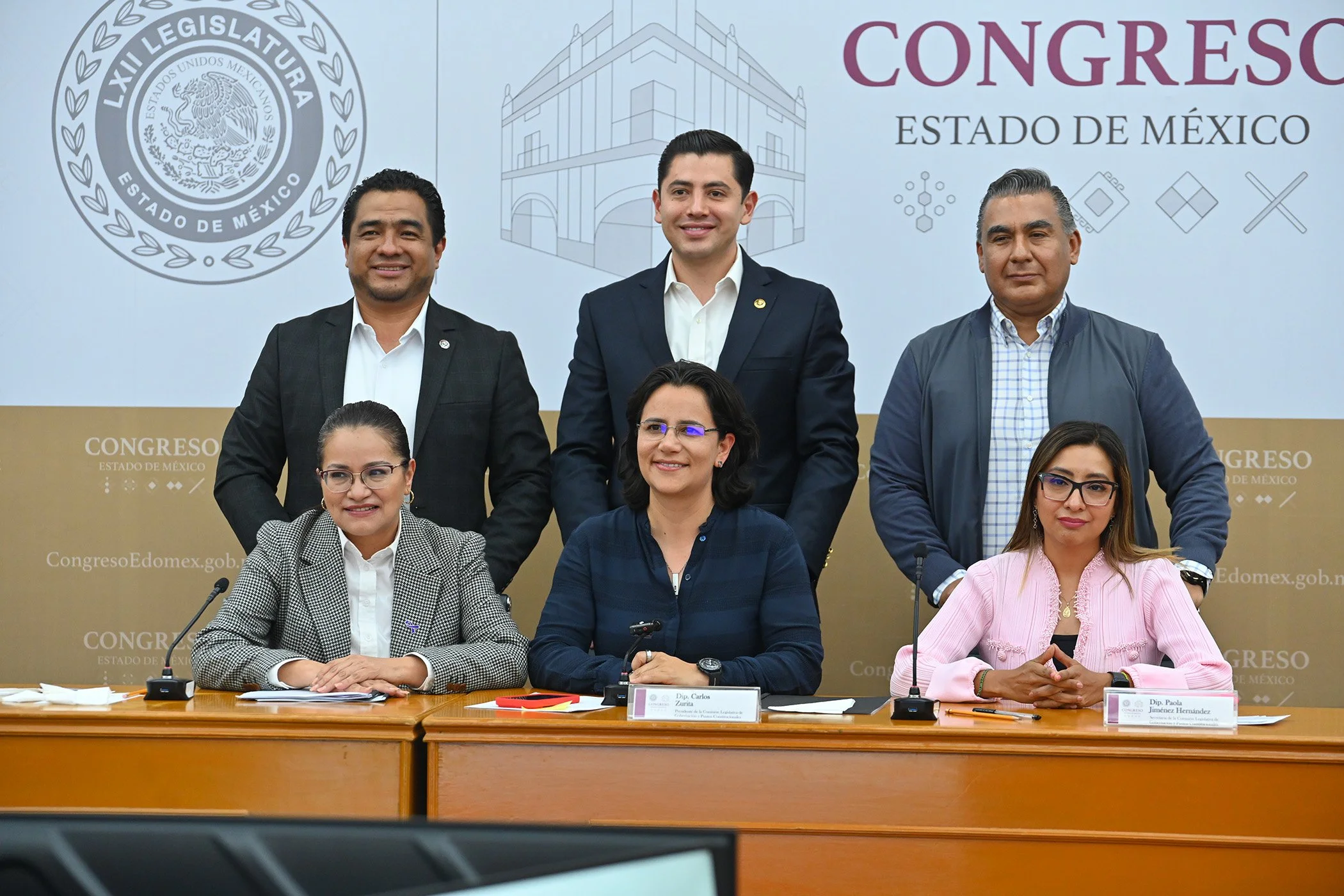 Group of six people, three men standing and three women seated at a table during a formal event or press conference, with a backdrop featuring government and legislative symbols and text.