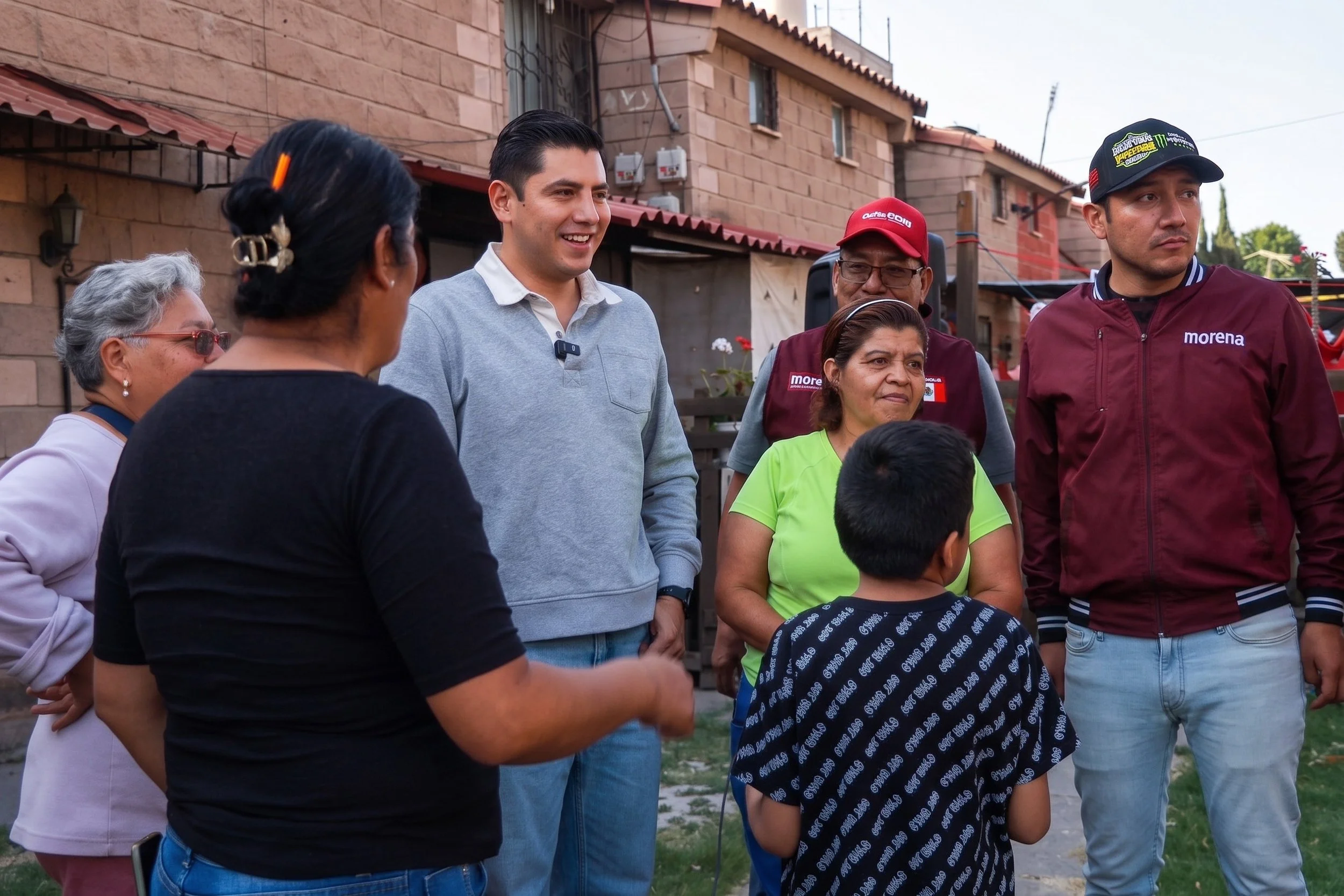 A group of people, including men and women of Hispanic or Latino descent, gathered outdoors in a neighborhood with brick houses in the background. They are engaged in conversation and appear to be smiling and listening attentively.