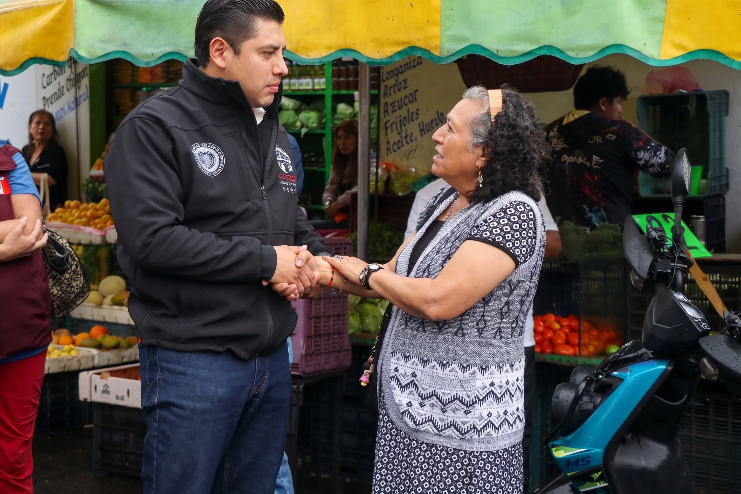 A young man and an older woman shake hands and converse at an outdoor market with fresh produce in the background.