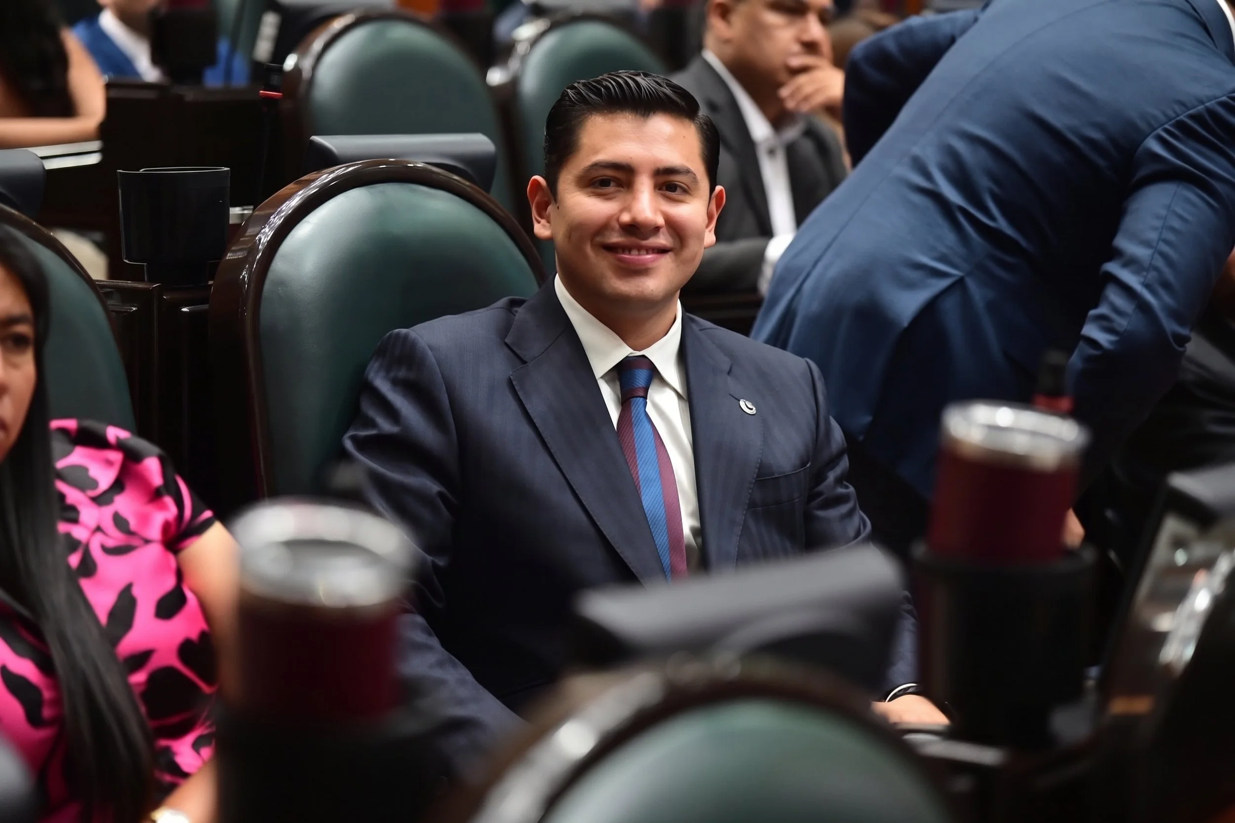 A man in a pinstripe suit with a pink and blue striped tie, sitting in a chamber with green seats, smiling at the camera, surrounded by other people.