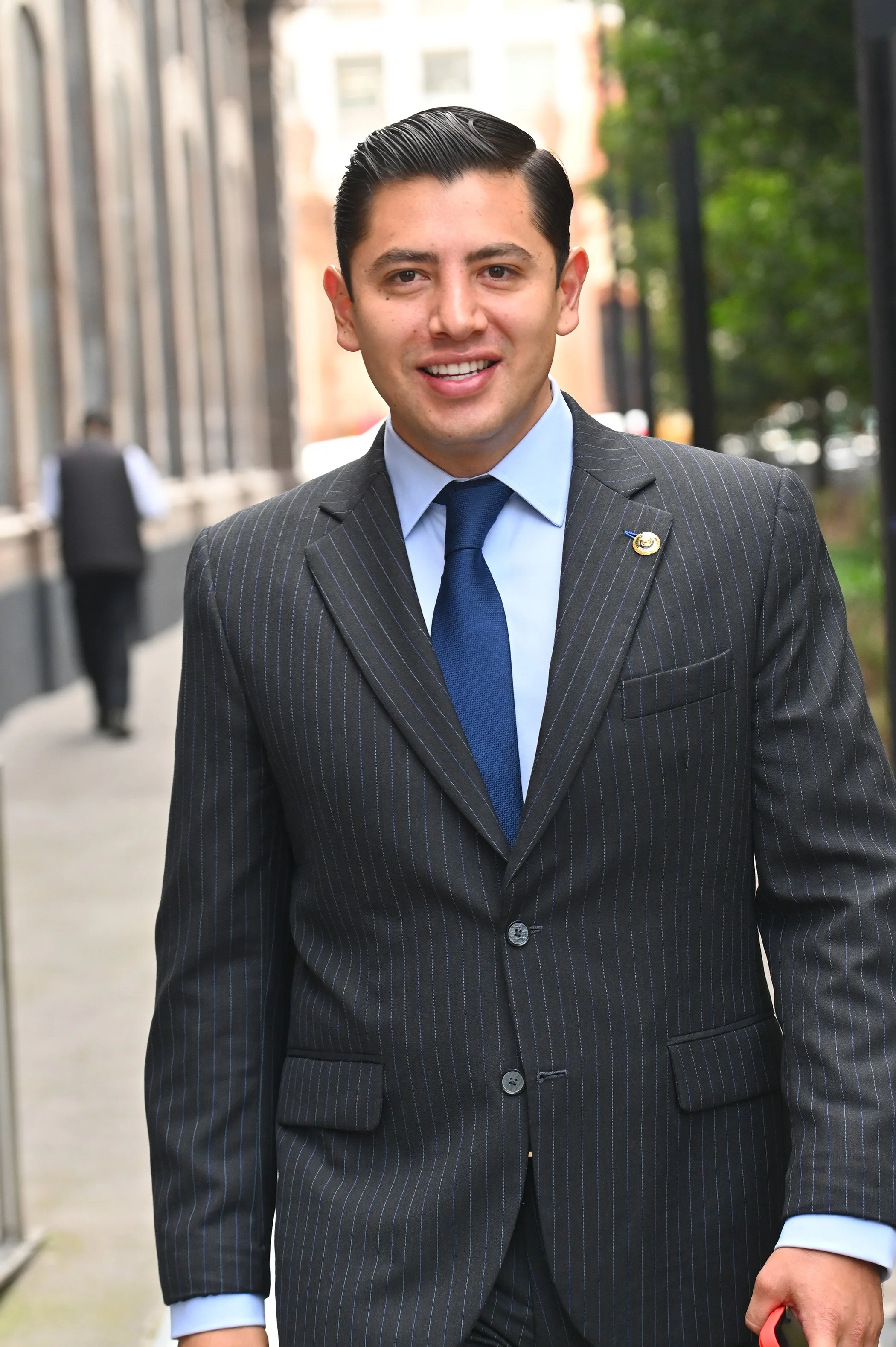 A young man in a dark pinstripe suit, blue shirt, and matching tie walking outdoors in an urban setting. He has slicked-back dark hair and is smiling, with a small pin on his lapel.