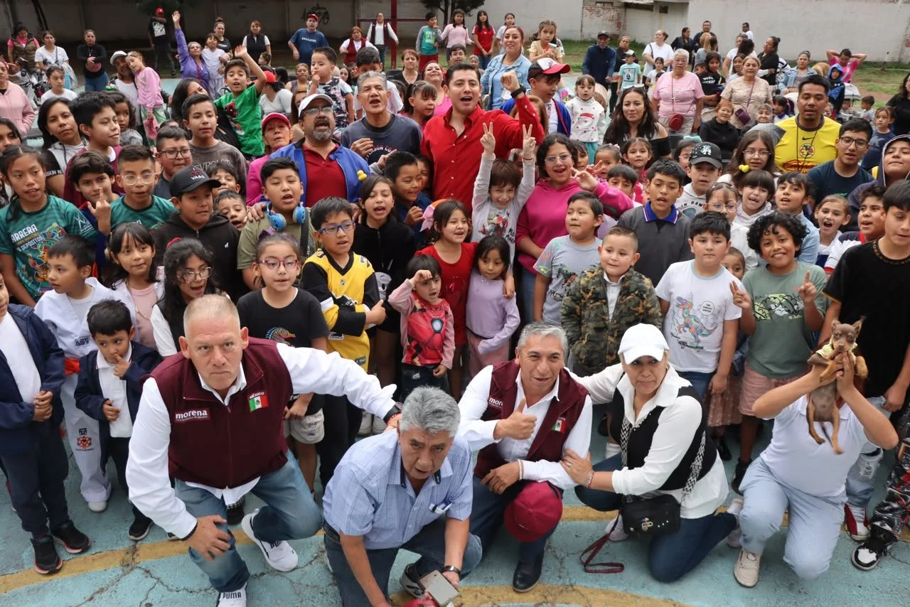 A large group of children and adults gathered outdoors, some smiling and making peace signs, with a few pets among them.