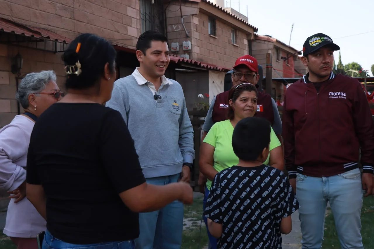 A group of seven people, including men and women, standing outdoors in a residential area and engaging in a conversation, with some smiling and appearing friendly.