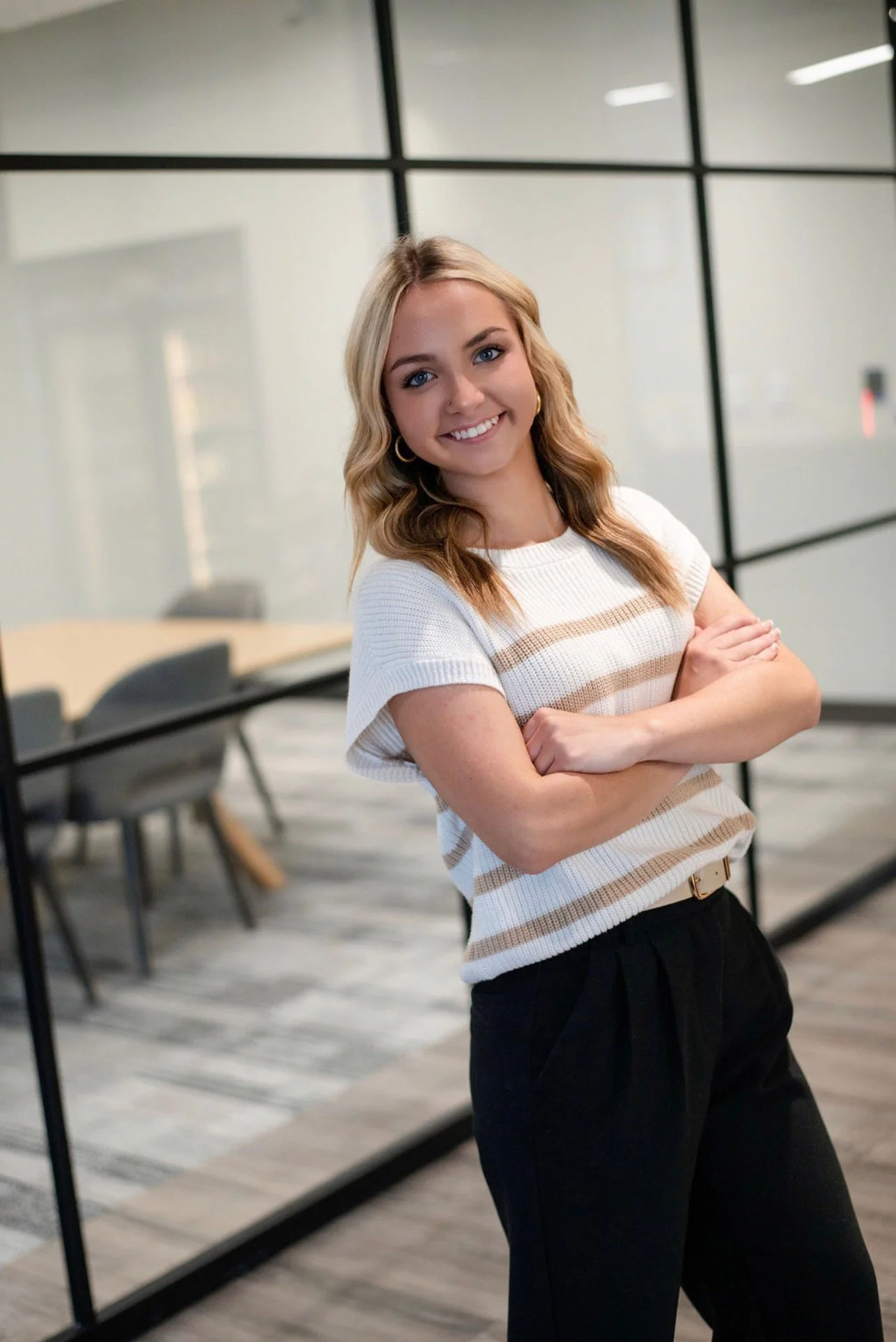 A young woman with blonde hair and blue eyes smiling with arms crossed in an office conference room with glass partitions and modern furniture.