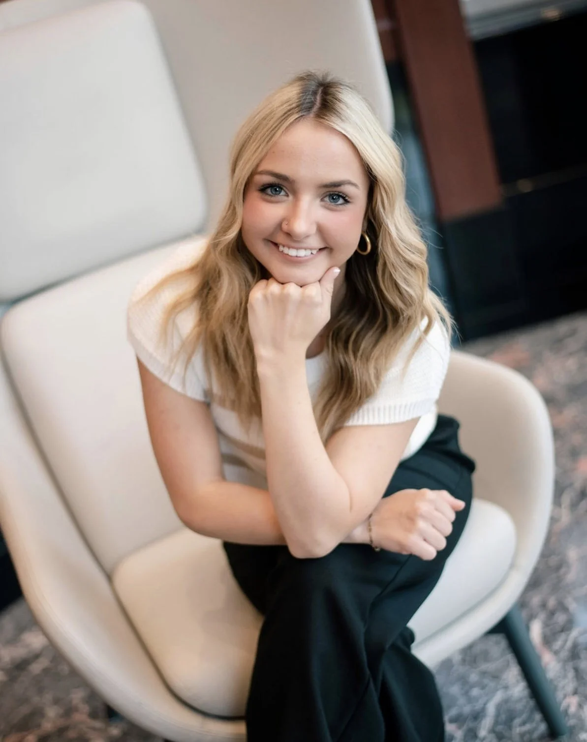 A young woman with blonde hair and blue eyes, smiling, sitting in an armchair with her chin resting on her hand.